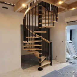 Nowoczesne schody spiralne z drewnianymi stopniami i czarną balustradą w jasnym wnętrzu. Widoczne elementy wykończeniowe i zabezpieczenia podłogi.