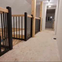 Wnętrze z drewnianymi schodami i czarną balustradą z pionowych prętów. Widoczne drewniane słupy i przygotowane podłoże. W tle oświetlone pomieszczenie.