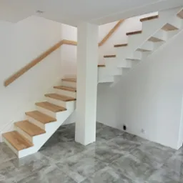 Nowoczesne schody z drewnianymi stopniami i białymi policzkami, bez balustrady, w jasnym wnętrzu z szarą podłogą. Minimalistyczny design, widok z dołu.