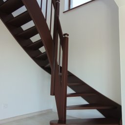 Usługi Stolarsko ślusarskie Franciszek Miszczak - Brązowe schody drewniane z prostą balustradą w jasnym wnętrzu. Widoczne stopnie, poręcze i fragment okna łukowego. Minimalistyczny design.