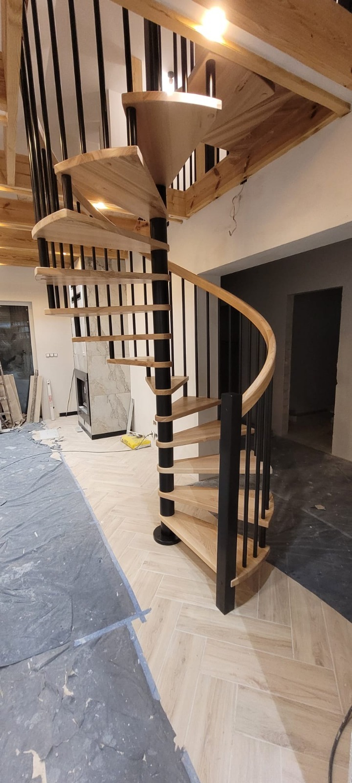 Spiralne schody z jasnego drewna z czarnymi metalowymi balustradami, widoczne na tle wnętrza w trakcie wykańczania, z podłogą w jodełkę i fragmentem zakrytej folią powierzchni.