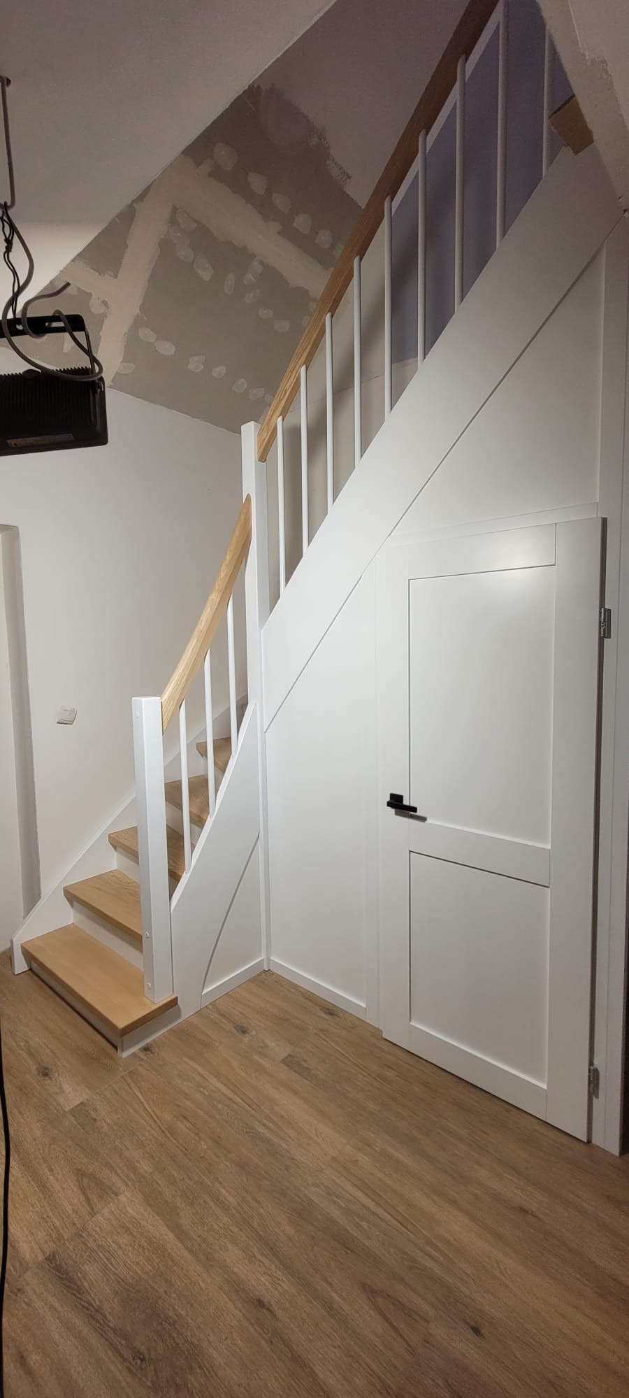 Jasne schody drewniane z białą balustradą i ukrytymi drzwiami pod schodami, na podłodze panele imitujące drewno. Na suficie widoczne ślady prac wykończeniowych.