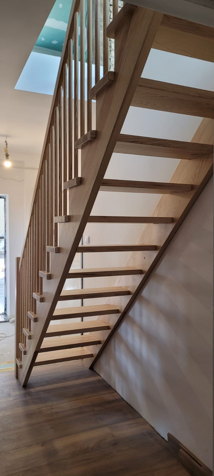 Jasne, drewniane schody z prostą balustradą w nowoczesnym wnętrzu. Widoczne drewniane stopnie i pionowe elementy balustrady. Na podłodze panele imitujące drewno.