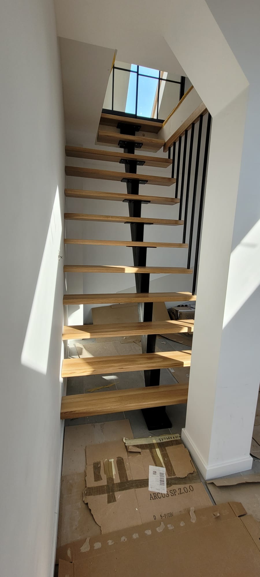 Nowoczesne schody drewniane z czarną metalową konstrukcją nośną. Stopnie z jasnego drewna, minimalistyczna balustrada. Widok z dołu, częściowo osłonięte podłoże.