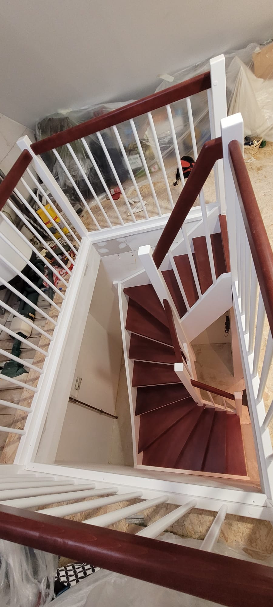 Wewnętrzne schody spiralne z drewnianymi stopniami w odcieniach brązu i białą balustradą, widziane z góry. Schody prowadzą w dół, oświetlone światłem z góry. Część pomieszczenia zabezpieczona folią.