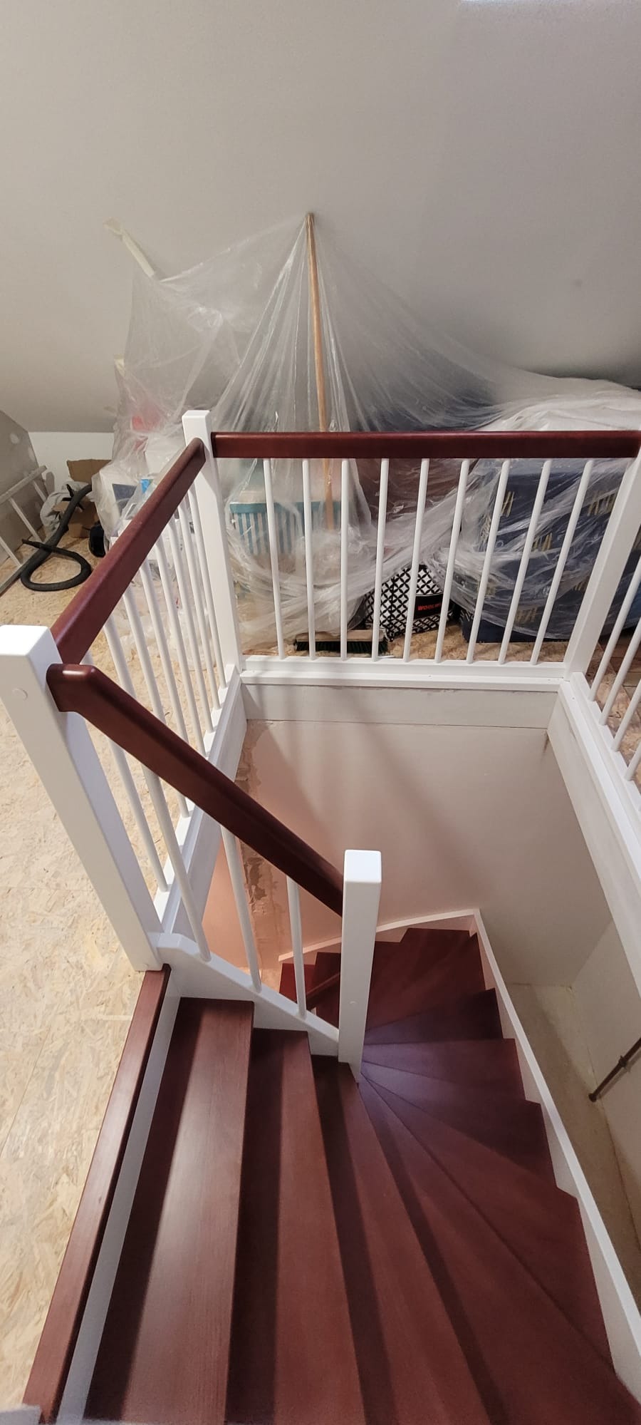 Widok z góry na spiralne schody z drewnianymi stopniami w odcieniu wiśni, białe balustrady i zabezpieczone folią otoczenie podczas remontu.