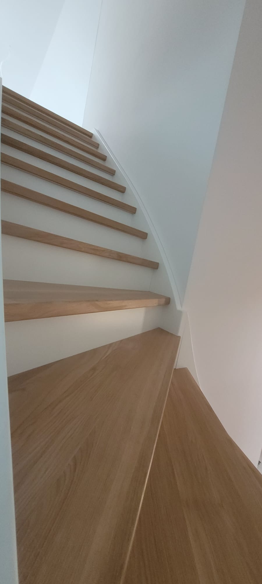 Minimalistyczne schody drewniane z jasnego drewna, w białym wnętrzu. Ujęcie od dołu podkreśla zakrzywiony kształt i nowoczesny design.