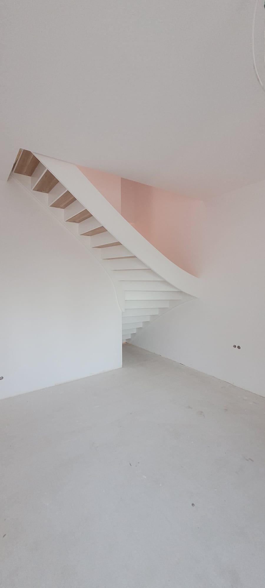 Minimalistyczne schody drewniane z białymi stopnicami i podstopnicami, zakręcające w lewo, w surowym, jasnym wnętrzu z różowym akcentem na ścianie.