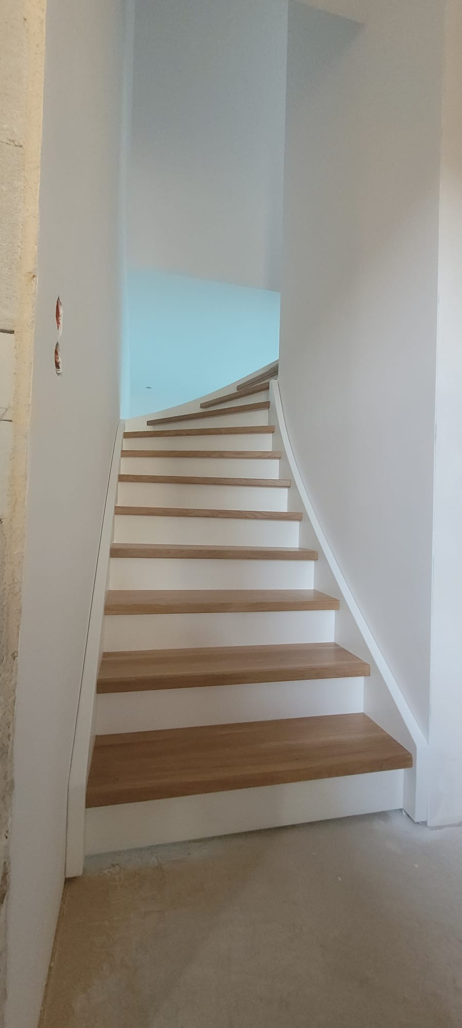 Nowoczesne schody z drewnianymi stopniami i białymi podstopnicami w jasnym wnętrzu. Widok z dołu na zakrzywiony bieg schodów.