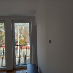 REHA-BUD SPÓŁKA Z OGRANICZONĄ ODPOWIEDZIALNOŚCIĄ - Wnętrze pokoju z balkonem w trakcie malowania, widoczna biała ściana z niedomalowanym fragmentem, grzejnik oraz fragment balkonu z czarną balustradą i widokiem na ulicę.