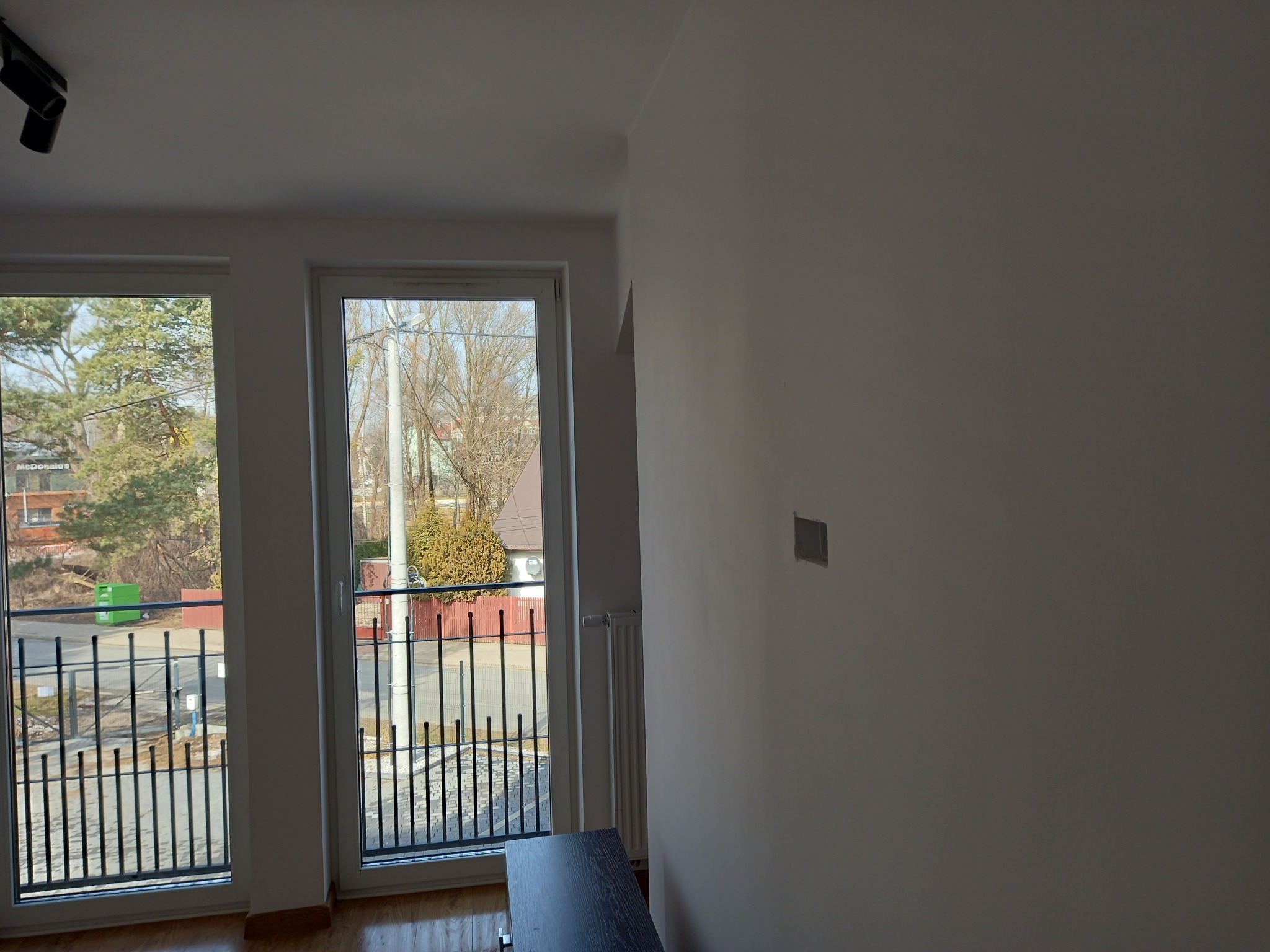 Wnętrze pokoju z balkonem w trakcie malowania, widoczna biała ściana z niedomalowanym fragmentem, grzejnik oraz fragment balkonu z czarną balustradą i widokiem na ulicę.