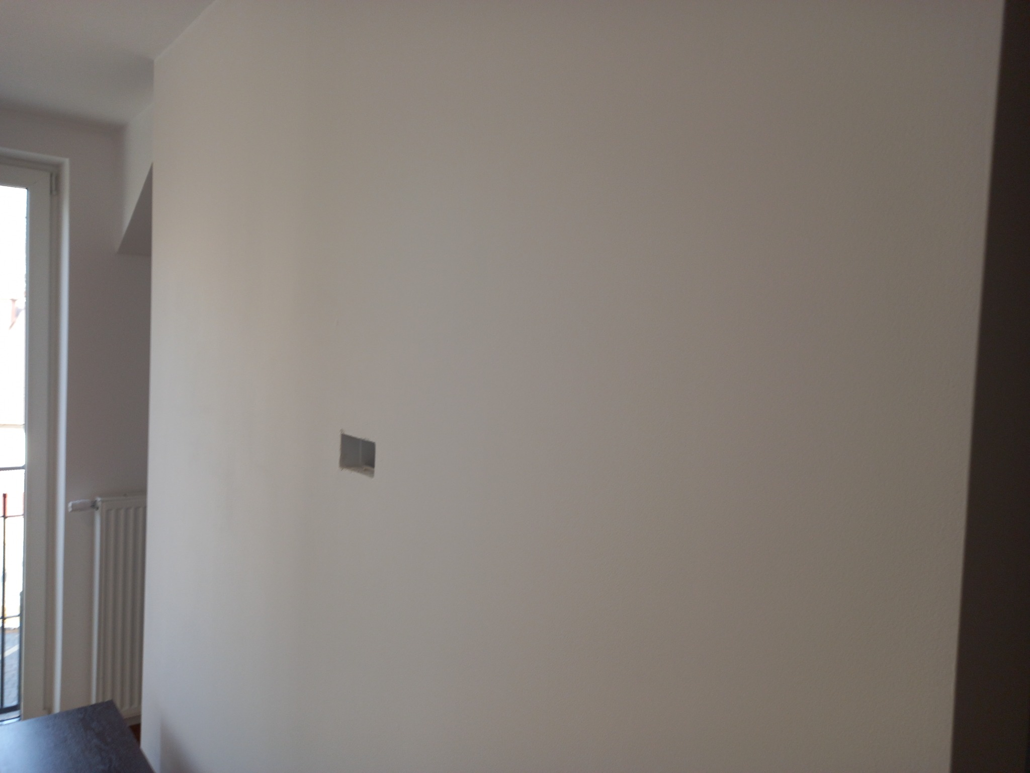 Świeżo pomalowana ściana w jasnym odcieniu z przygotowanym otworem na gniazdko elektryczne, widoczny fragment grzejnika i okna.