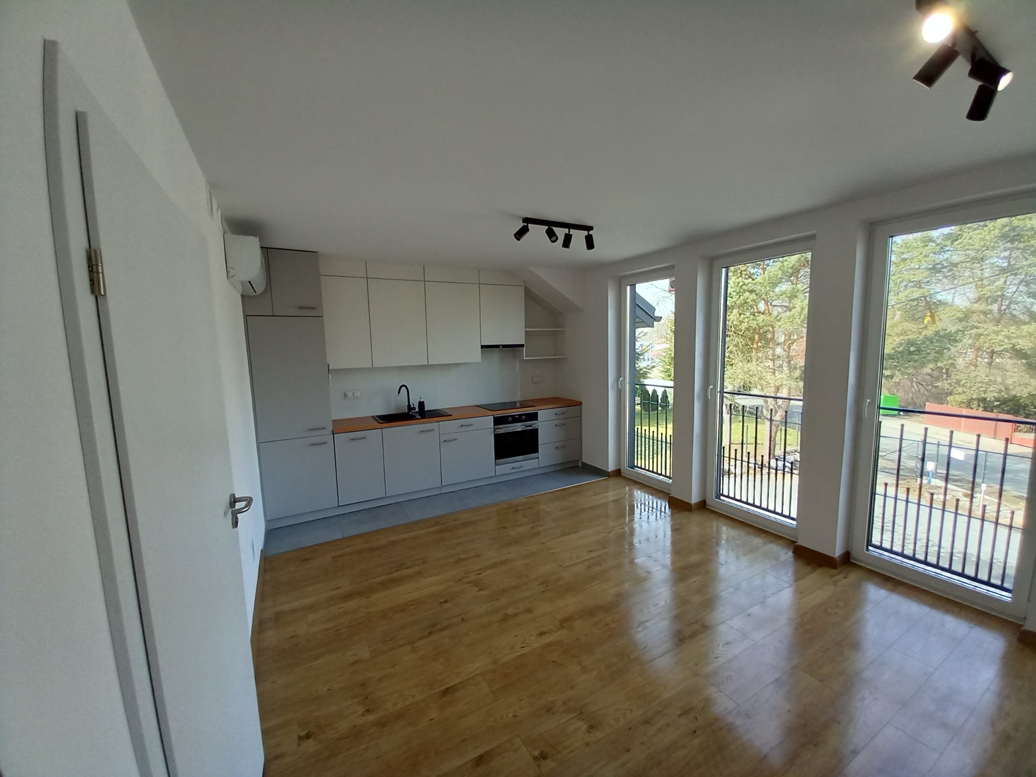 Jasne, przestronne wnętrze w Krakowie z drewnianą podłogą, nowoczesną zabudową kuchenną w odcieniach szarości i bieli, dużymi oknami balkonowymi z widokiem na zieleń oraz minimalistycznym...