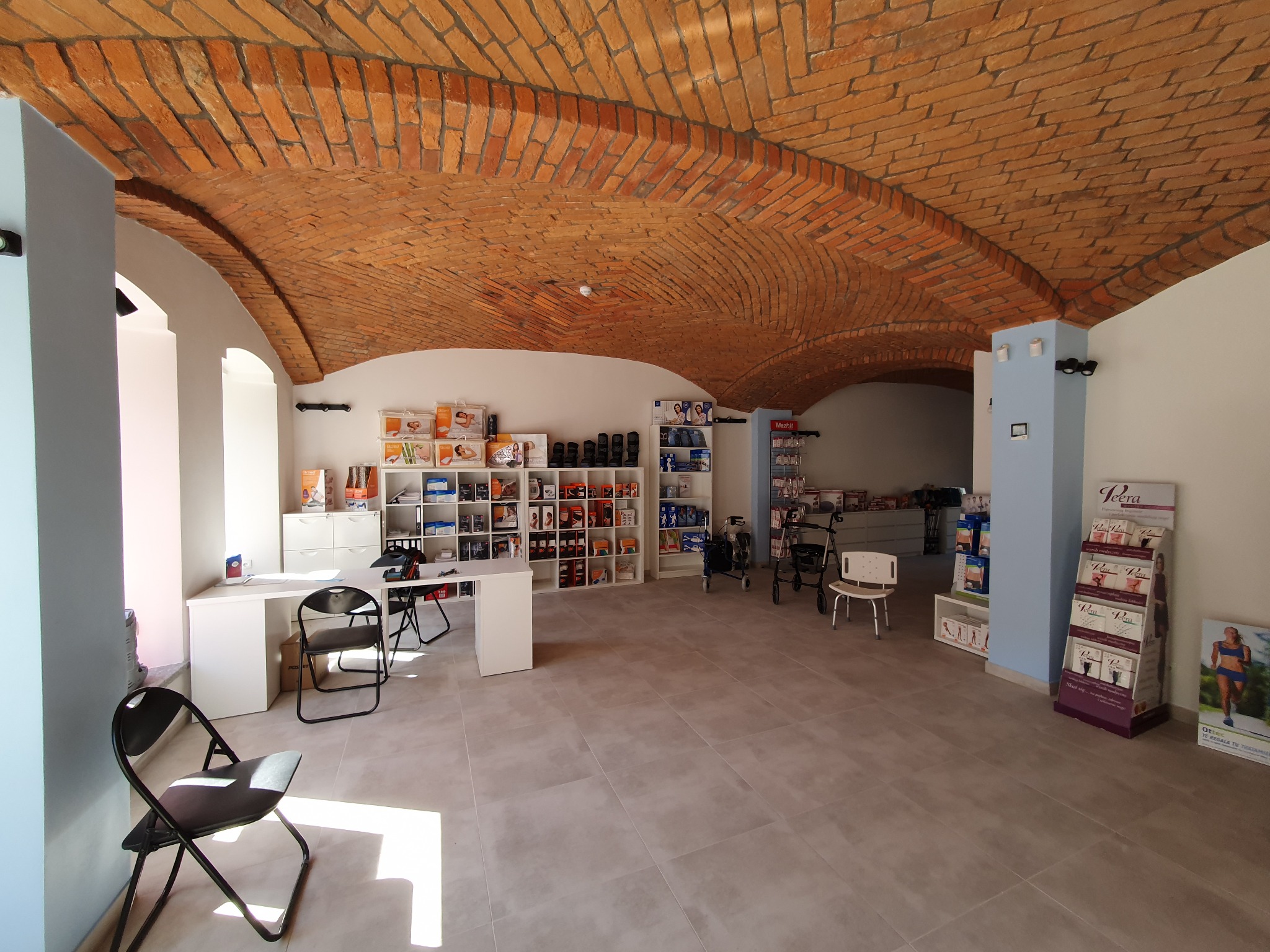 Jasne wnętrze biura z ceglanym sklepieniem łukowym, białymi ścianami, szarymi płytkami podłogowymi, białymi meblami i czarnymi krzesłami, częściowo wypełnione produktami na półkach.