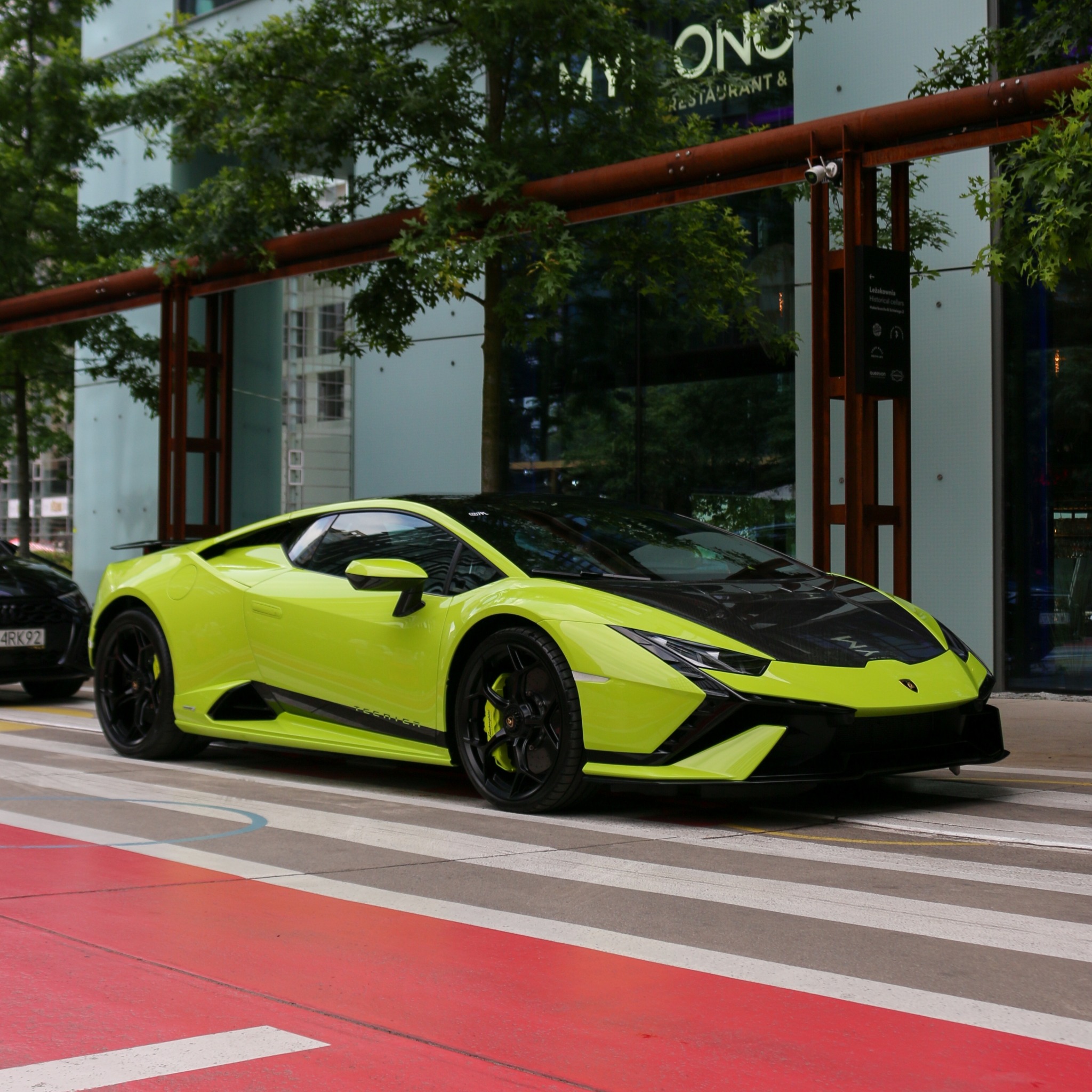 Neonowo-zielony Lamborghini Huracan z czarnym dachem i maską parkuje na czerwono-białym pasie dla pieszych przed nowoczesnym budynkiem z restauracją.