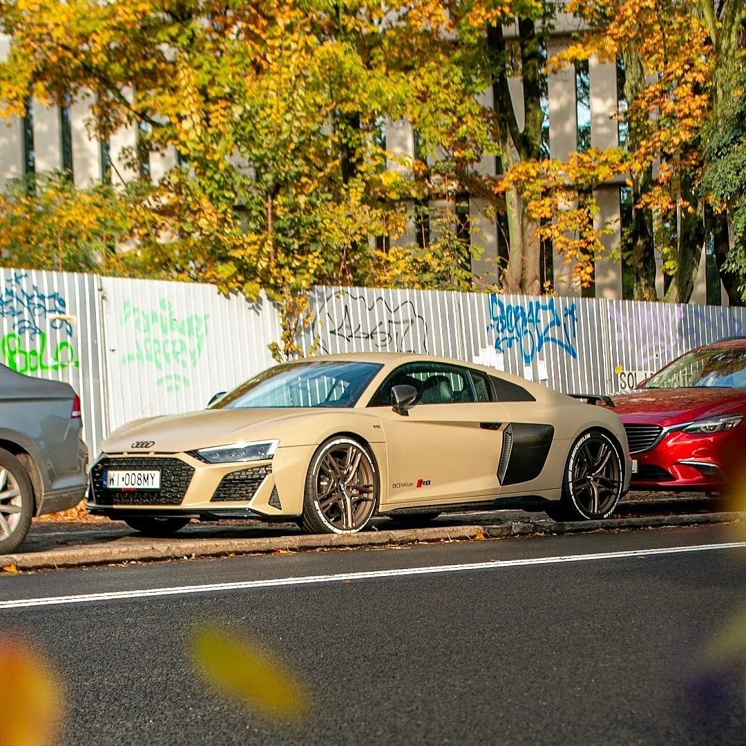 Piaskowy Audi R8 zaparkowany na ulicy z graffiti w tle, z widocznymi oponami z białymi napisami i tablicą rejestracyjną WI 008MY.