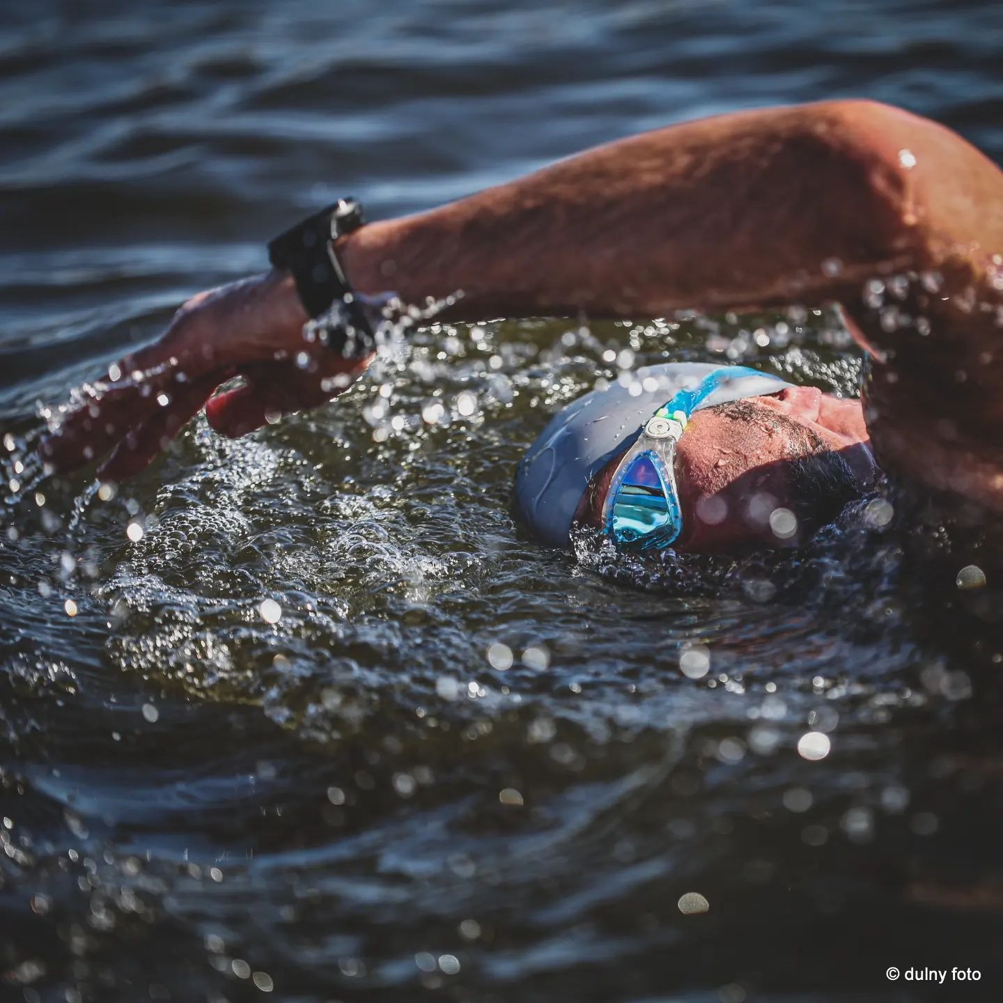 Pływający mężczyzna w czepku i okularach, woda spryskuje jego ramię podczas kraula, widoczny zegarek sportowy.
