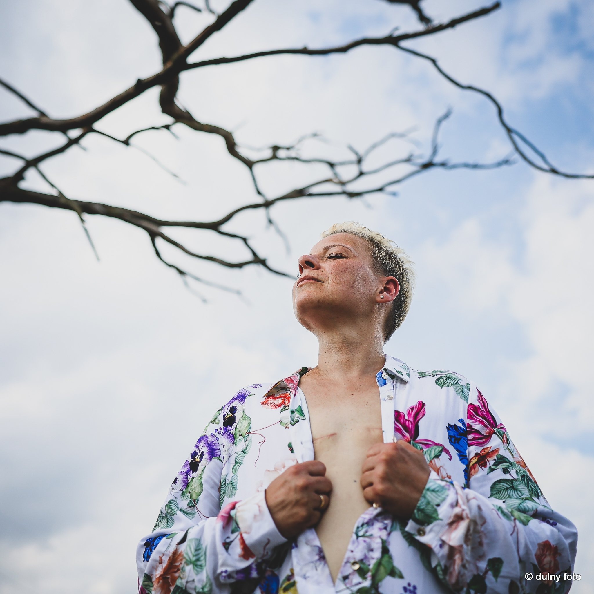Portret kobiety z krótkimi blond włosami, w kwiecistej koszuli, spoglądającej w górę na tle błękitnego nieba i gałęzi drzewa.