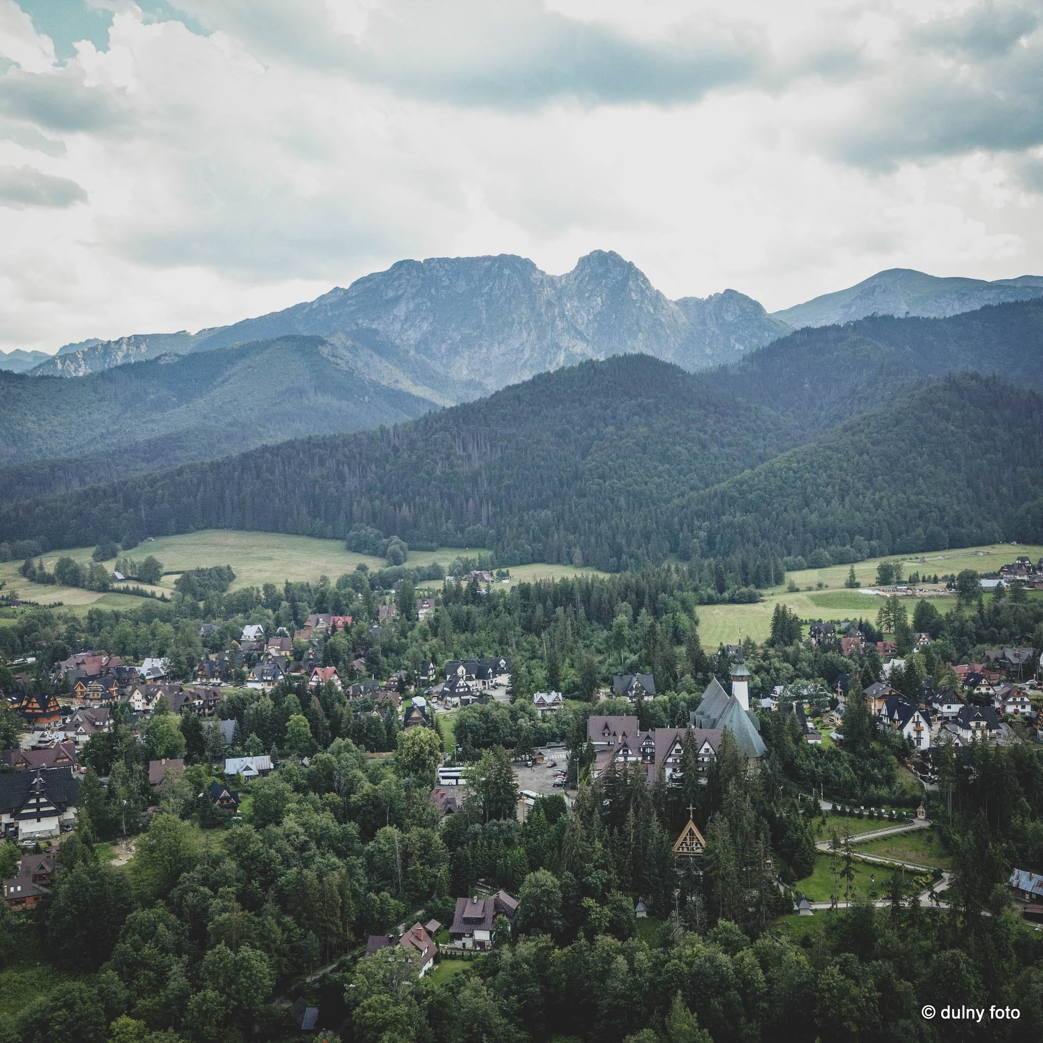 Panoramiczne ujęcie Zakopanego z widokiem na Giewont, wykonane z lotu ptaka, ukazuje architekturę podhalańską w otoczeniu lasów i gór.