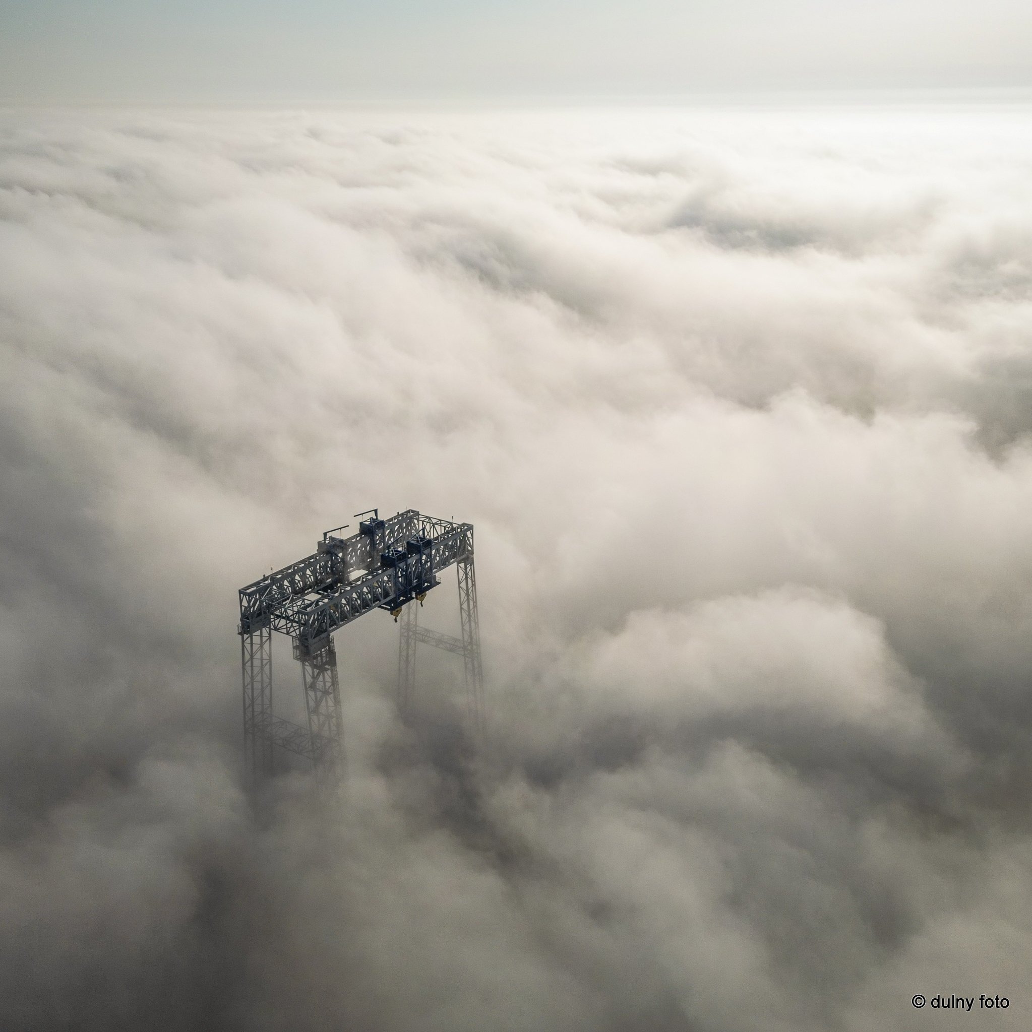 Metalowa konstrukcja dźwigu wyłaniająca się z gęstej warstwy mgły, widok z lotu ptaka.