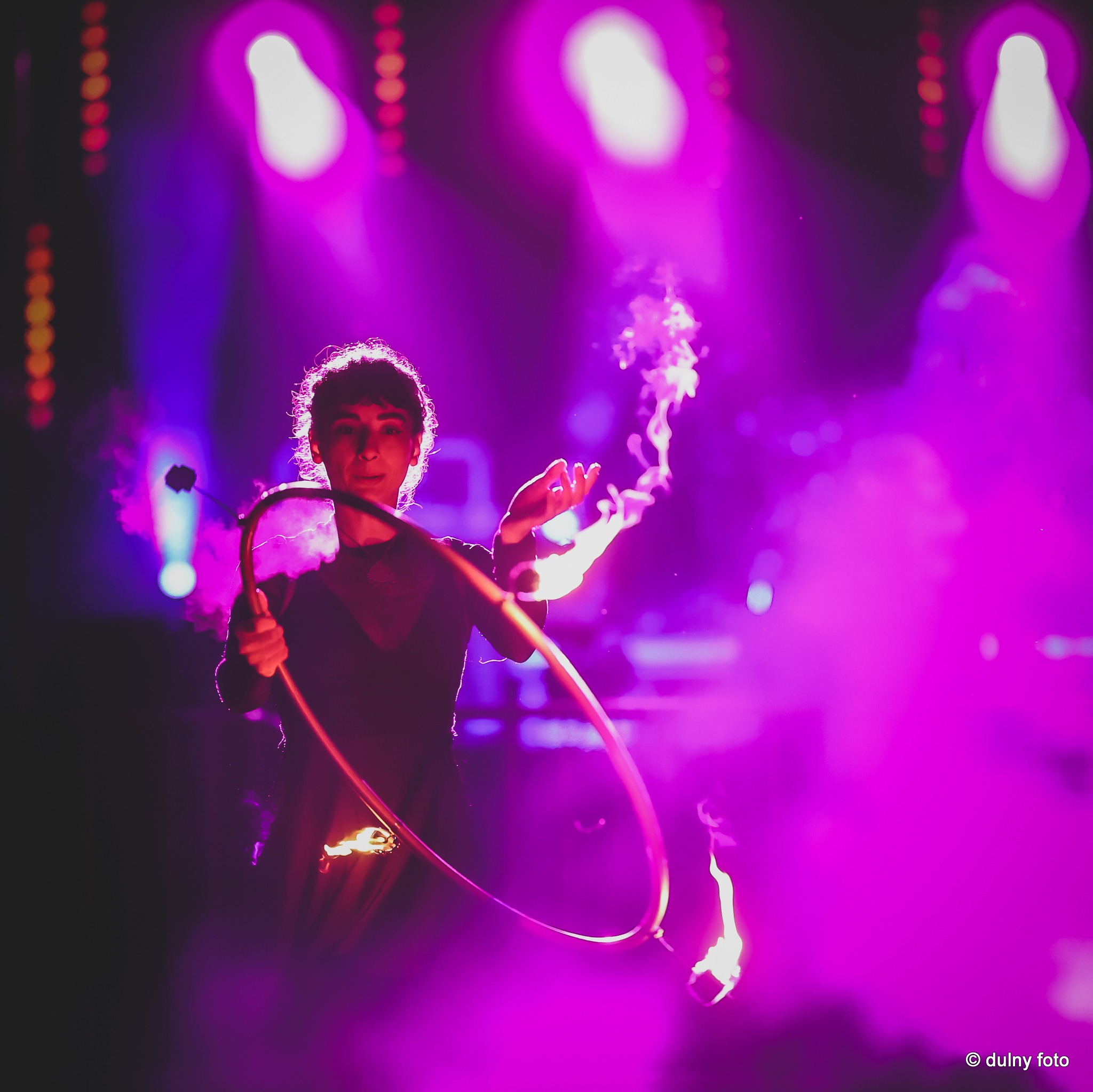 Kobieta wykonuje pokaz z ogniem, trzymając płonące rekwizyty na tle fioletowego oświetlenia scenicznego.