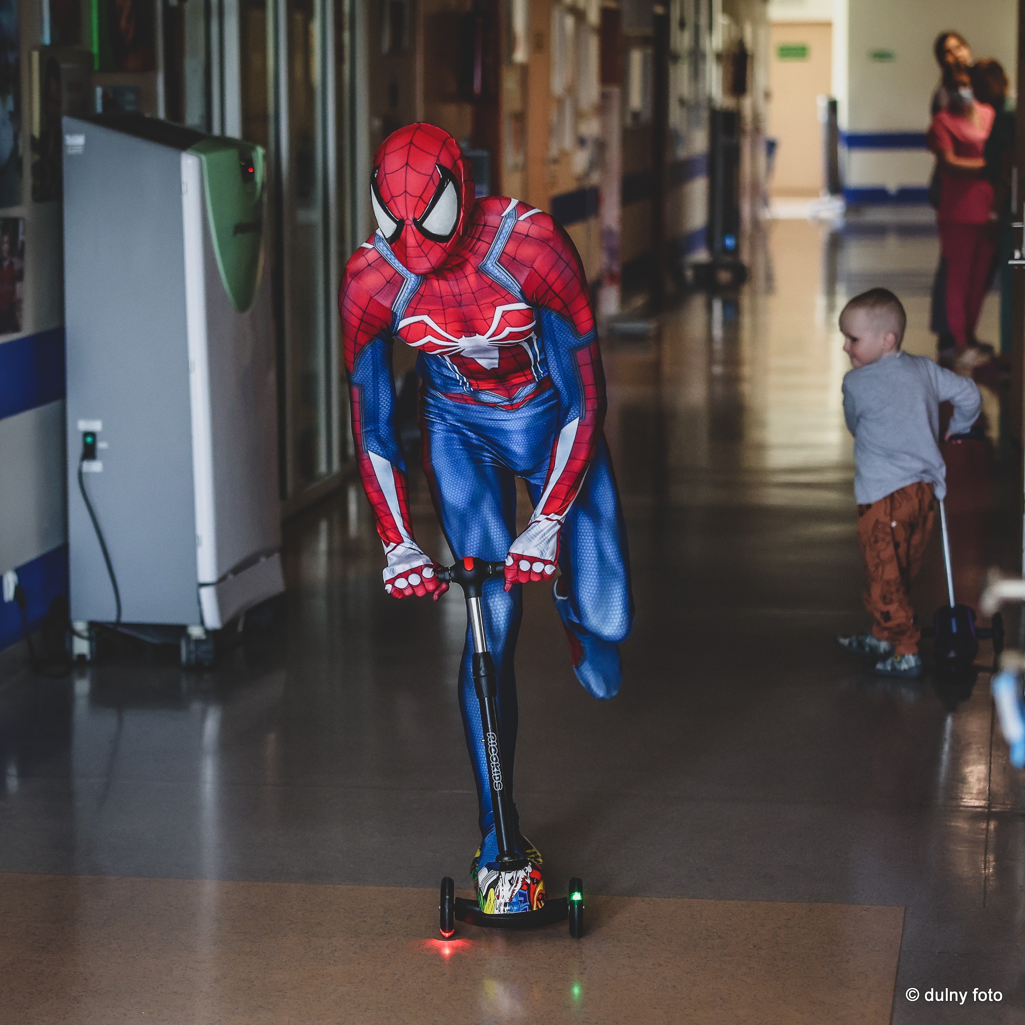 Osoba w kostiumie Spider-Mana na hulajnodze w szpitalnym korytarzu, w tle dziecko i dwie kobiety