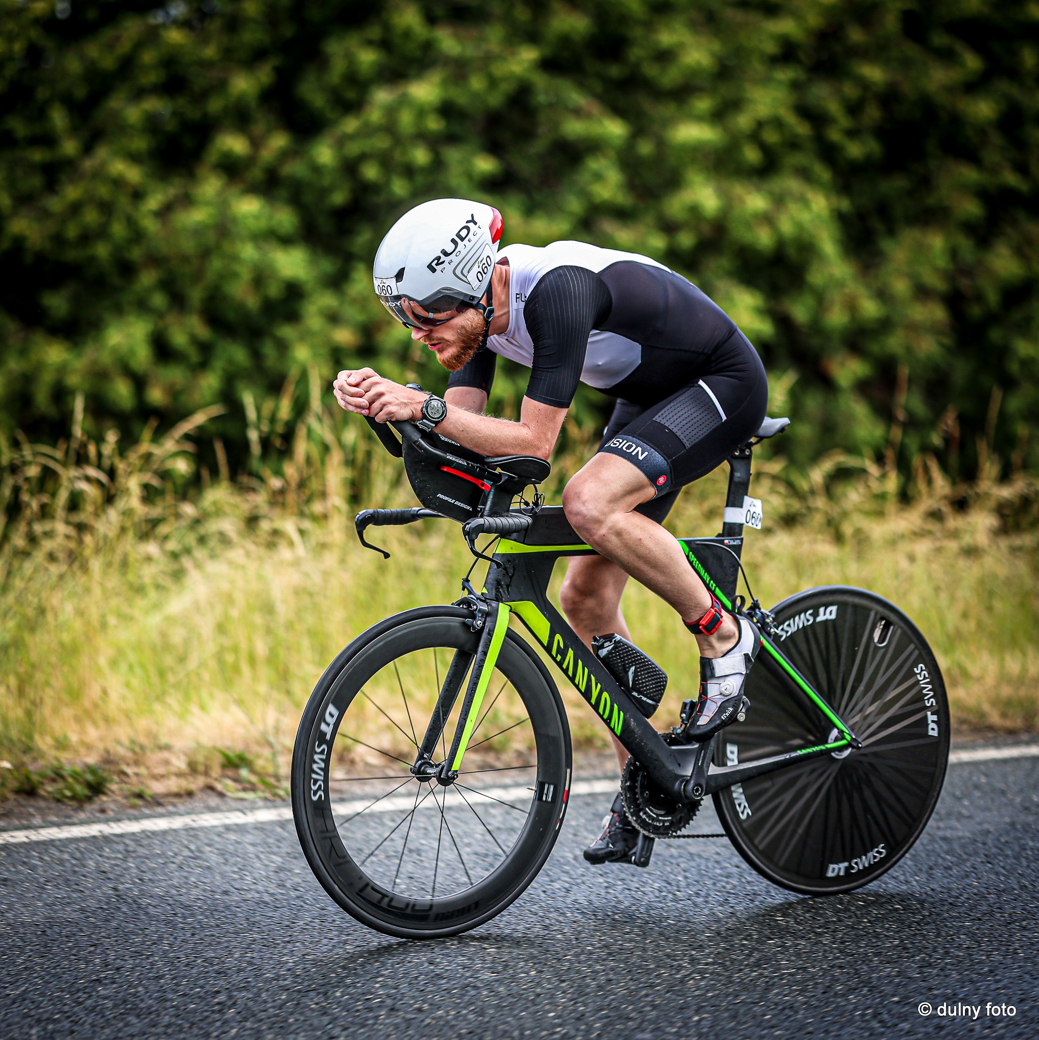 Mężczyzna w kasku i stroju triathlonowym na rowerze Canyon, z numerem startowym 060, podczas zawodów na asfalcie, w tle zieleń drzew.