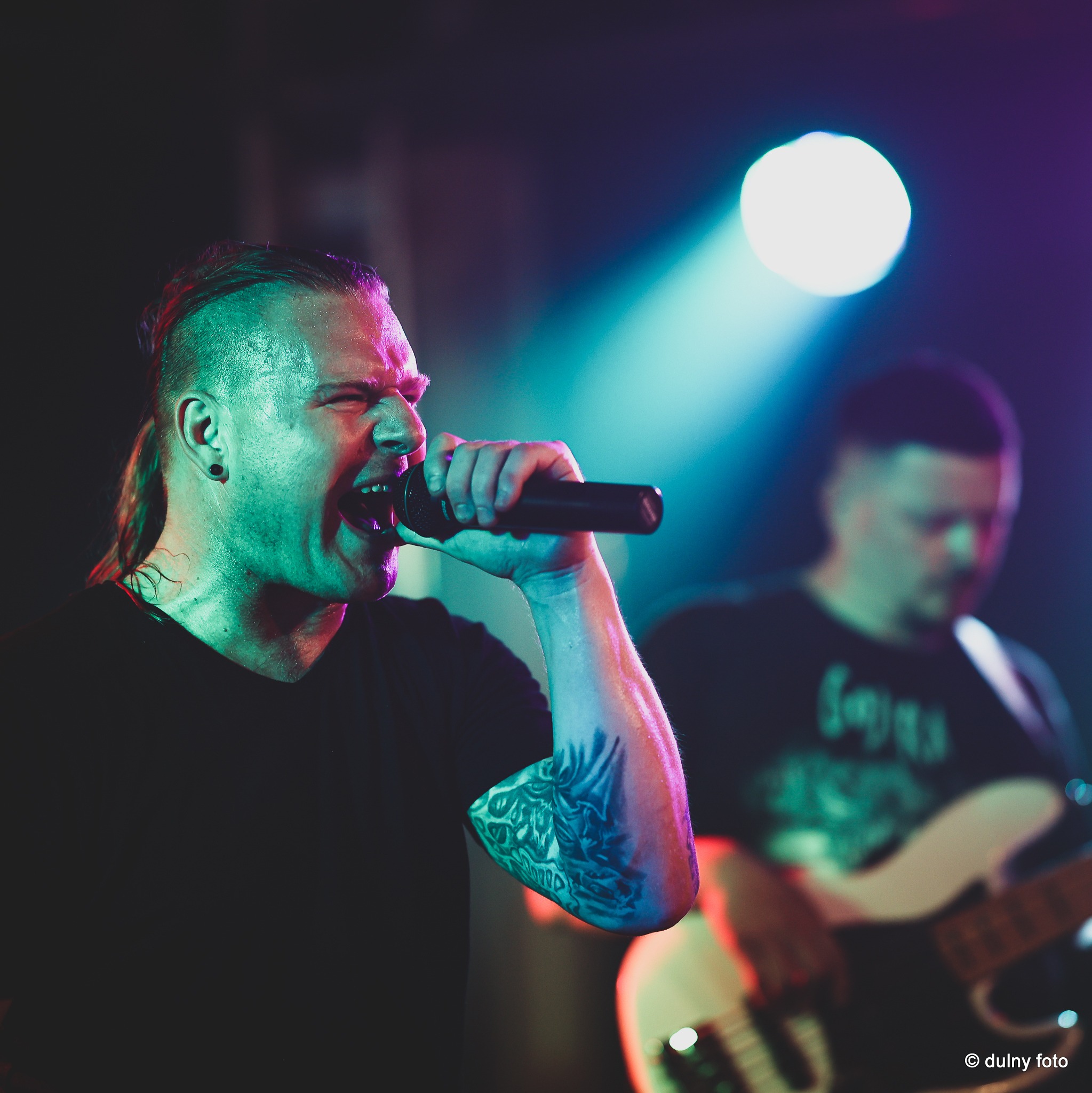 Wokalista z irokezem i tatuażem na ramieniu śpiewa do mikrofonu na scenie, oświetlony kolorowymi światłami, w tle basista.