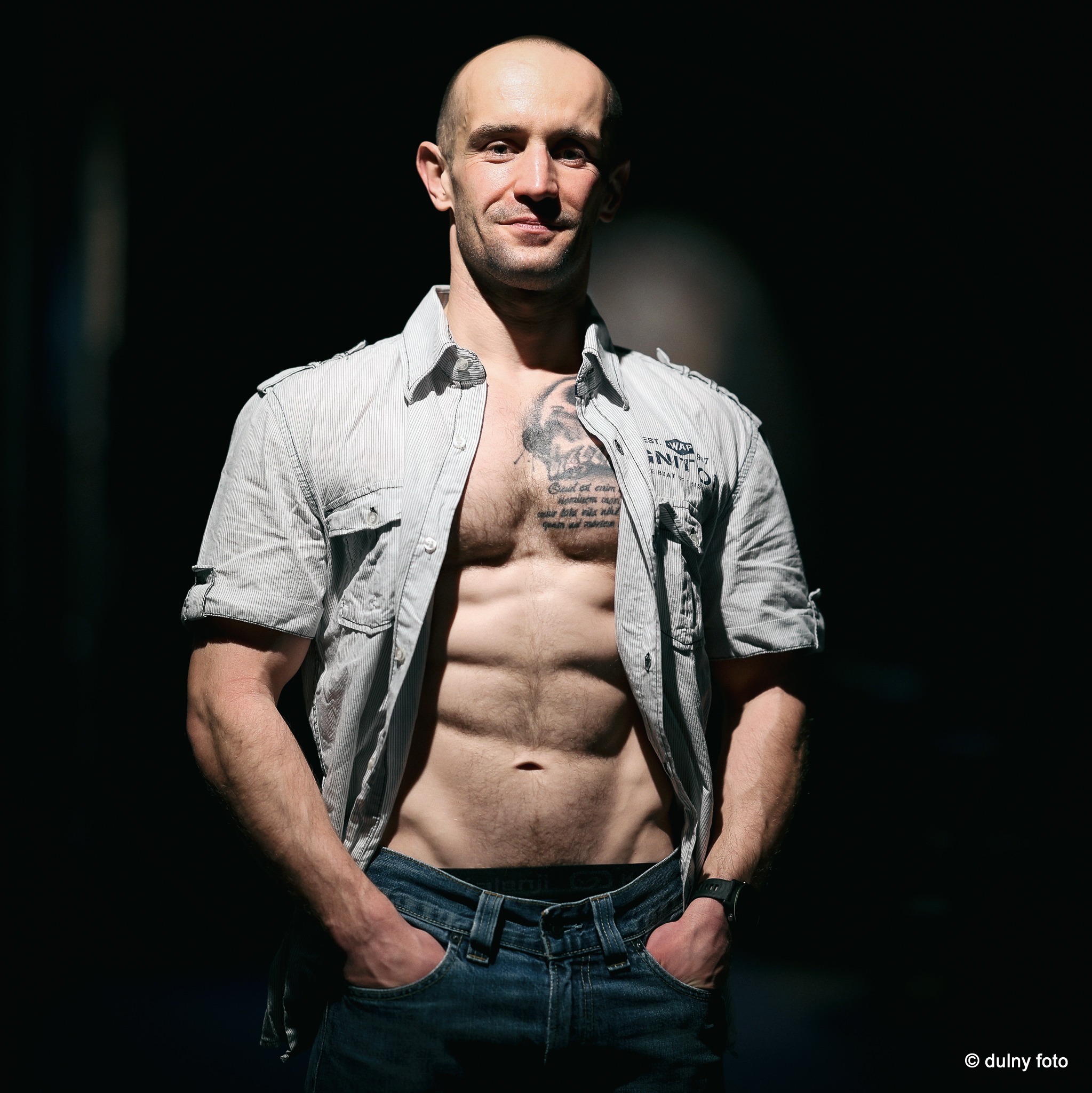 Mężczyzna z tatuażem na klatce piersiowej, w rozpiętej koszuli w paski i jeansach, pozujący w mocnym kontraście światła i cienia.