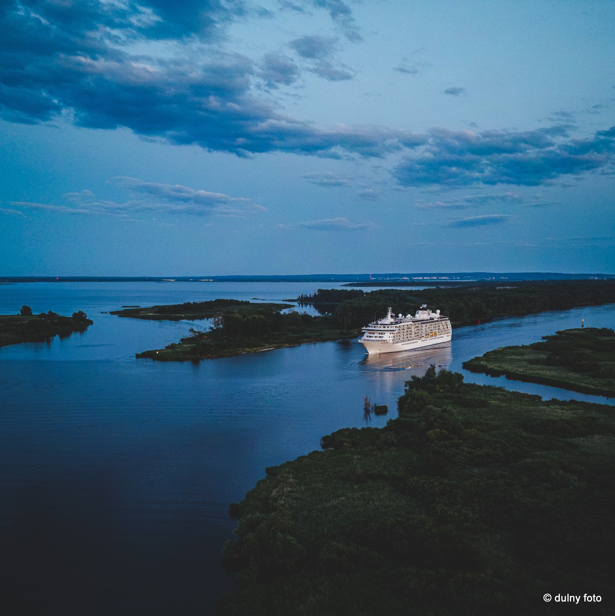 Duży, biały statek wycieczkowy płynie rzeką otoczoną zielenią, pod błękitnym niebem z ciemnymi chmurami, widziany z lotu ptaka.