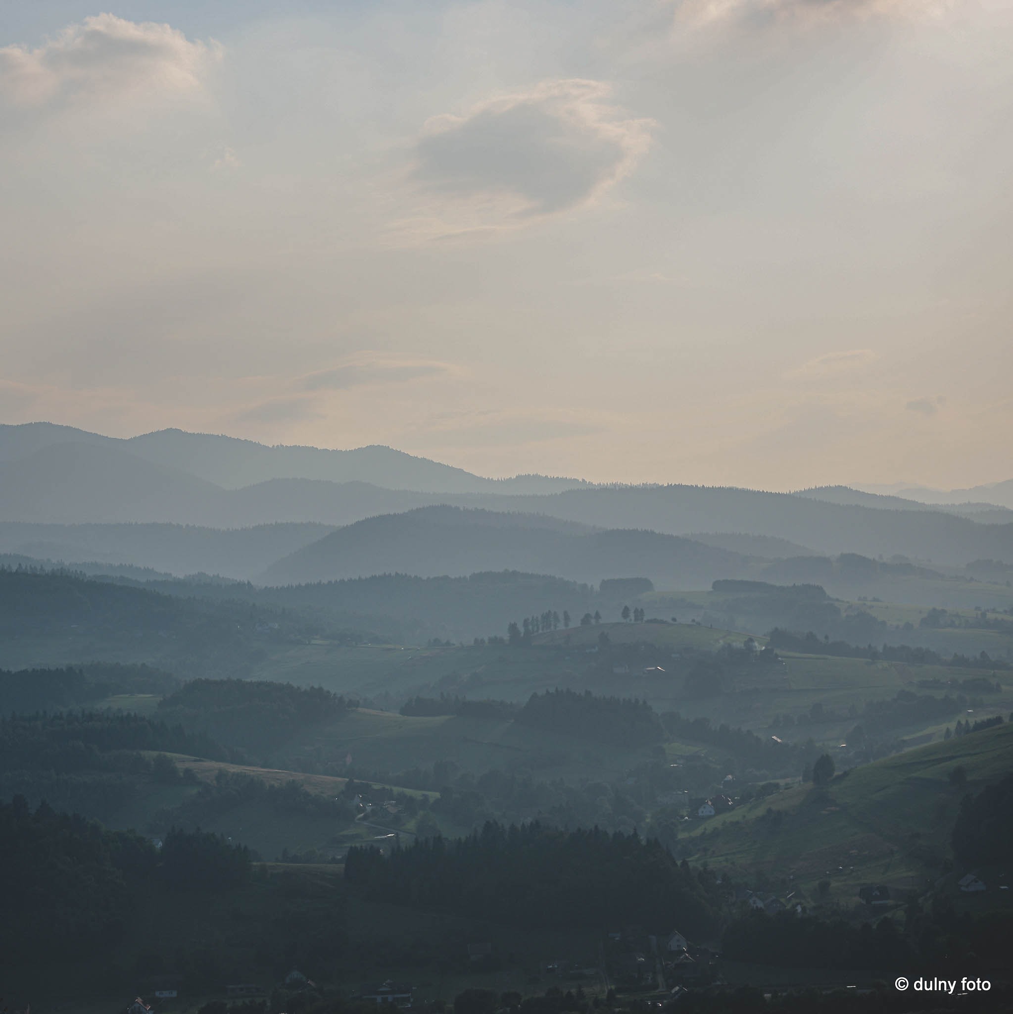 Panoramiczny widok na zalesione wzgórza i doliny spowite delikatną mgłą, z subtelnym światłem słonecznym przebijającym się przez chmury.