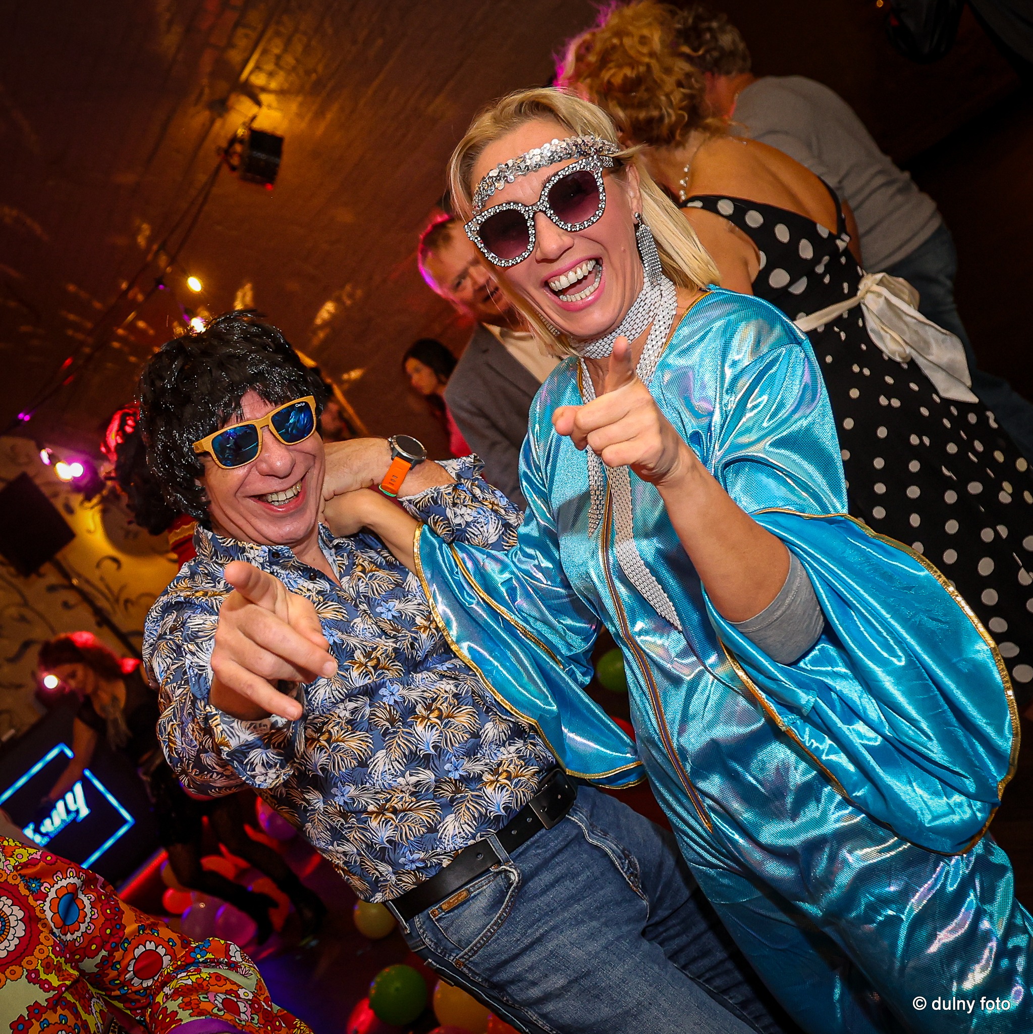 Rozbawiona para w kostiumach disco, z uśmiechem wskazująca palcami w Szczecinie, na tle innych osób i dekoracji imprezowych.