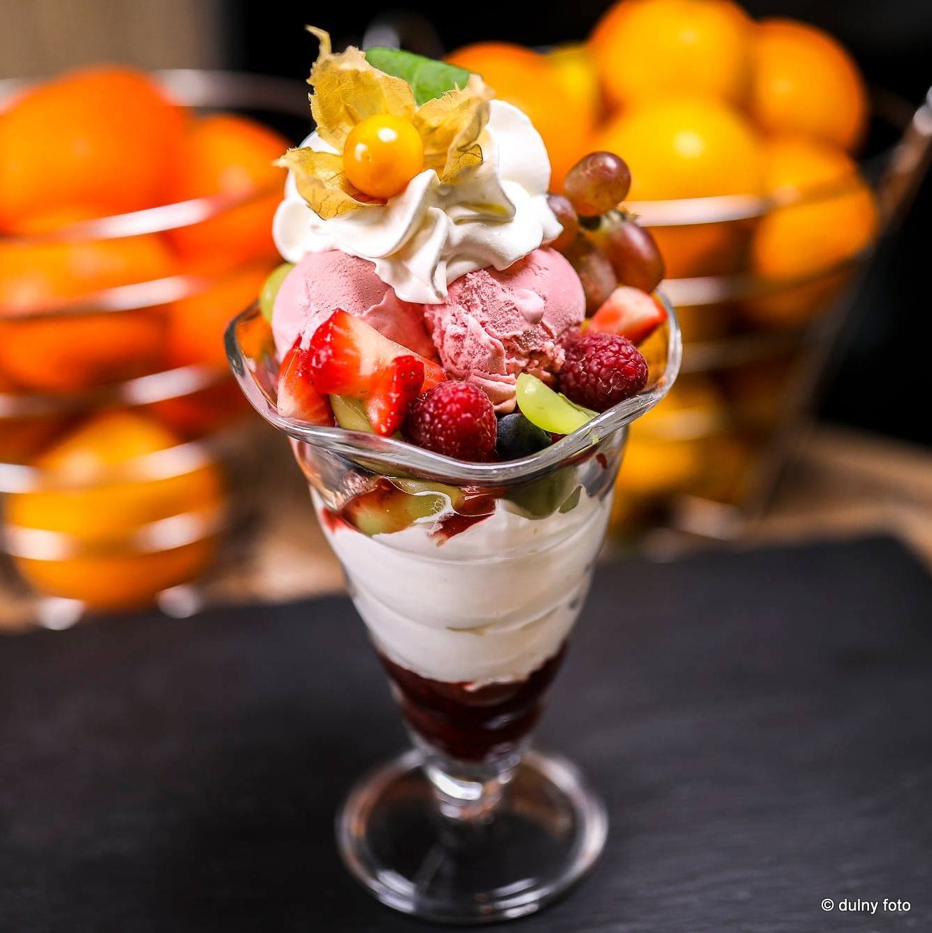 Deser lodowy w szklanym pucharku, udekorowany bitą śmietaną, physalis, malinami, truskawkami i zielonymi winogronami, na czarnym tle, z koszem pomarańczy w tle.