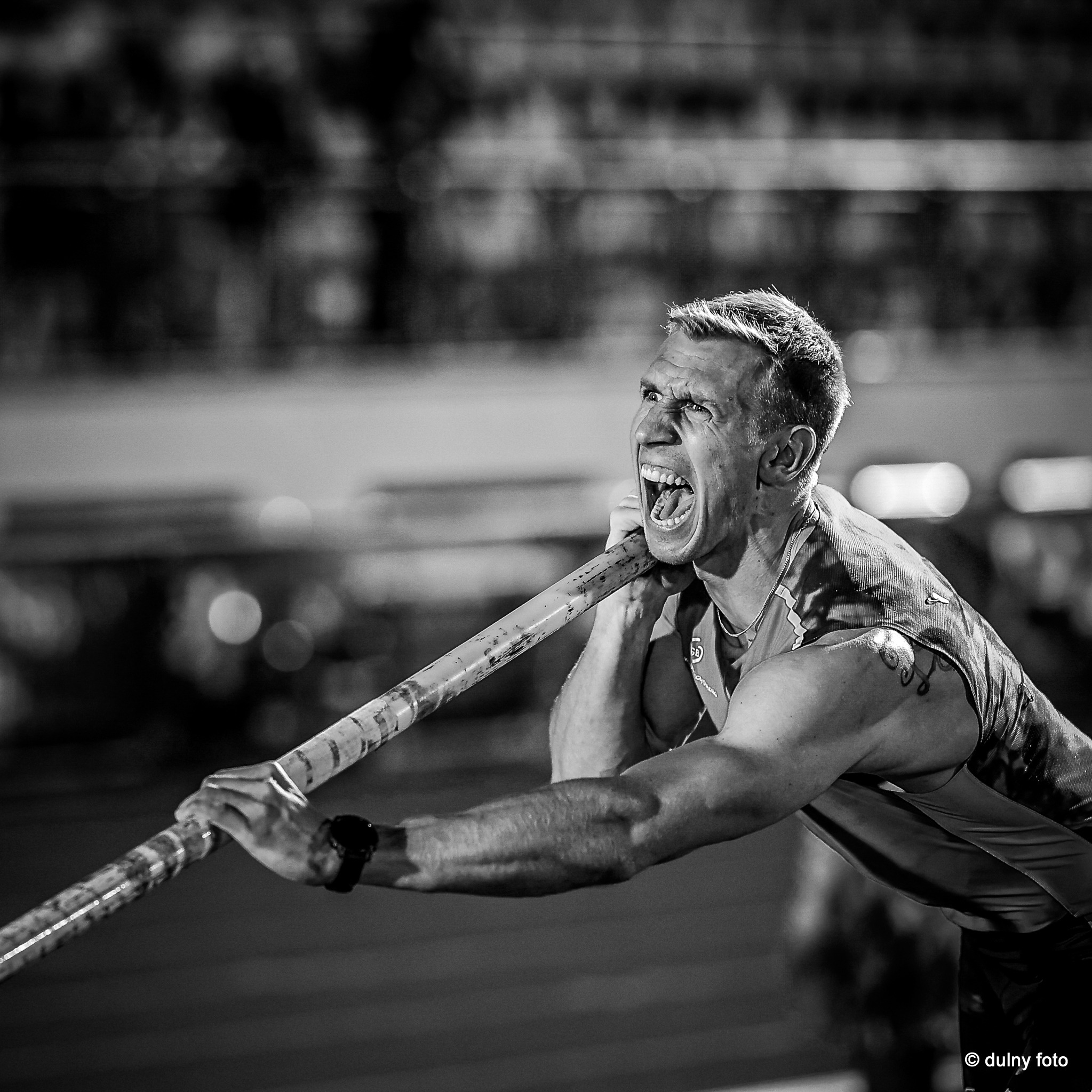 Czarno-białe zdjęcie sportowca krzyczącego z wysiłku podczas skoku o tyczce, widoczny tatuaż na ramieniu i zegarek na nadgarstku.