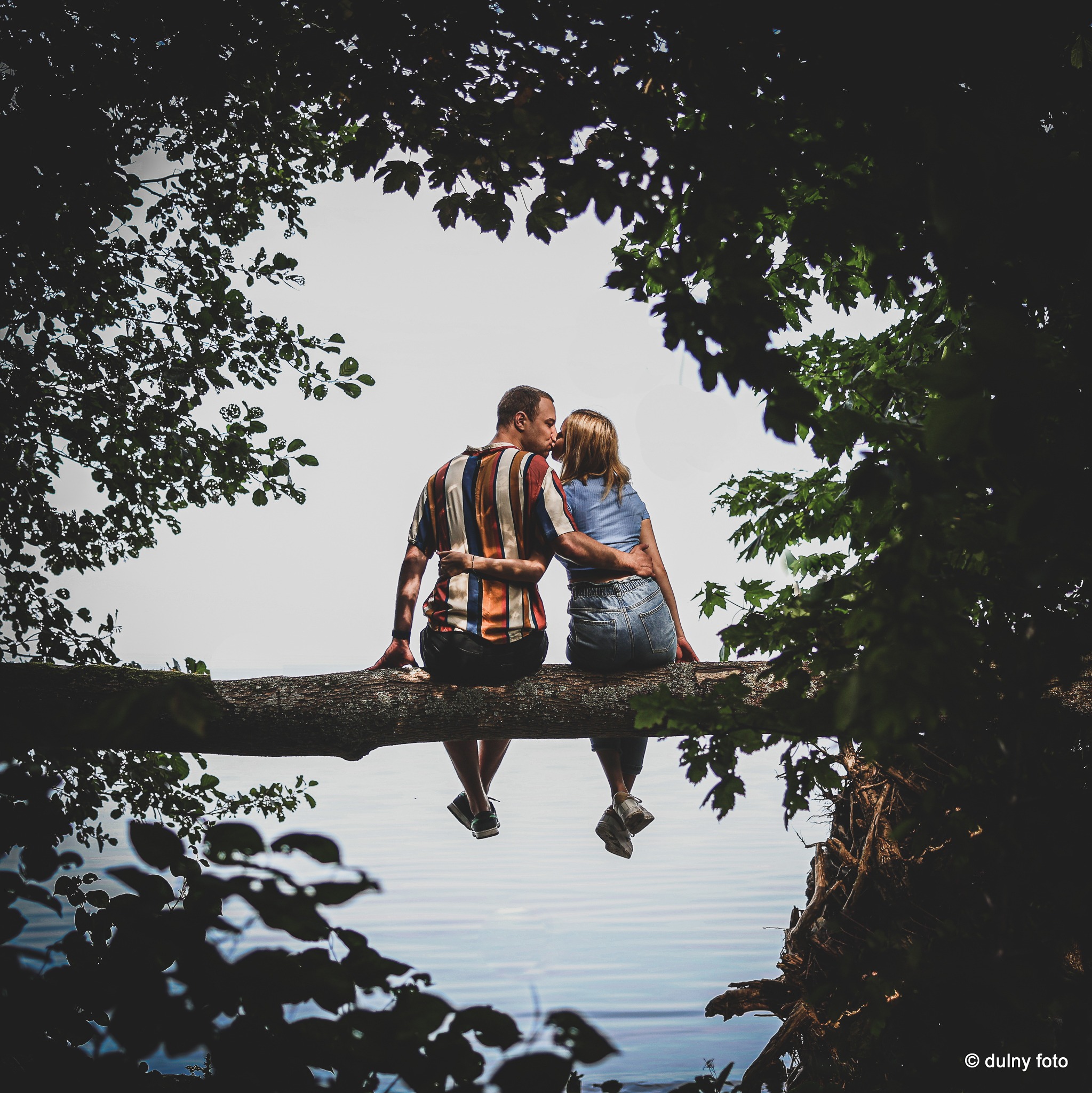 Para całująca się siedząc na powalonym drzewie z widokiem na wodę, otoczona liśćmi