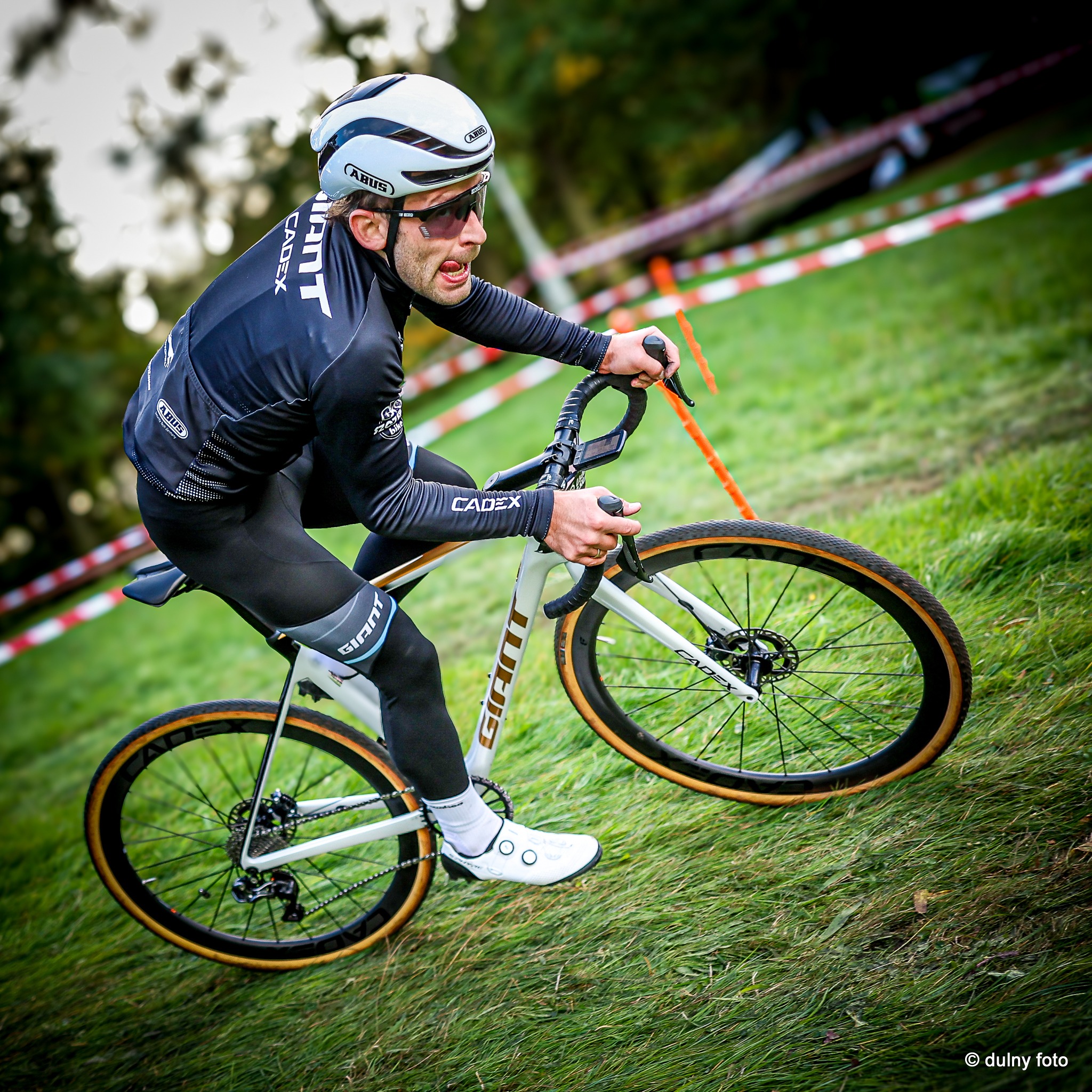 Kolarz w czarnym stroju i białym kasku marki ABUS, jadący na białym rowerze Giant Cadex po zielonej, trawiastej nawierzchni, na trasie wyznaczonej taśmami.