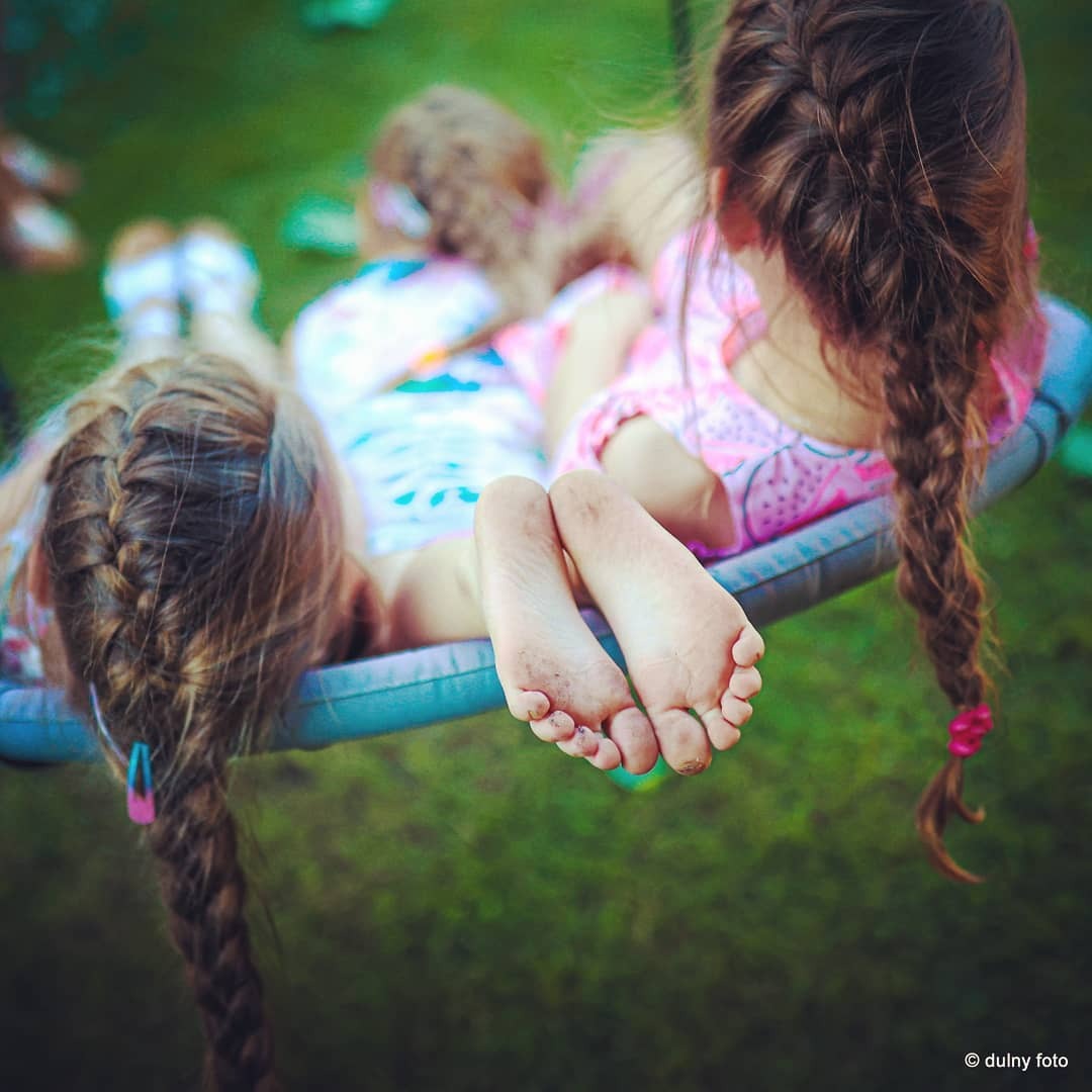 Trzy dziewczynki leżące na huśtawce, widoczne bose stopy jednej z nich z ziemią, zaplecione warkocze z kolorowymi gumkami na końcach, zielona trawa w tle.