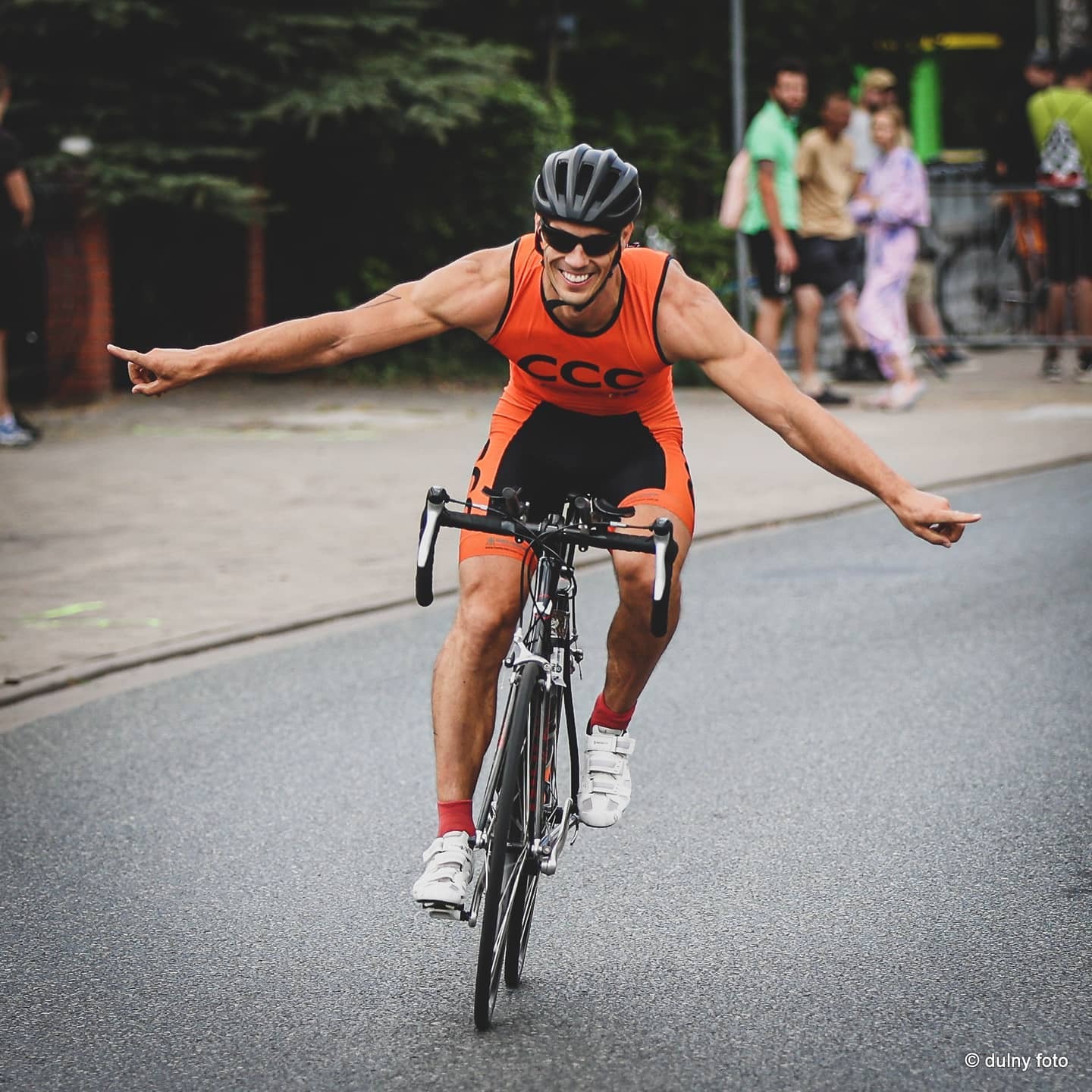 Uśmiechnięty triathlonista w pomarańczowo-czarnym stroju, kasku i okularach przeciwsłonecznych, jedzie na rowerze, wskazując palcami w dwie strony, na tle rozmytych widzów i zieleni.