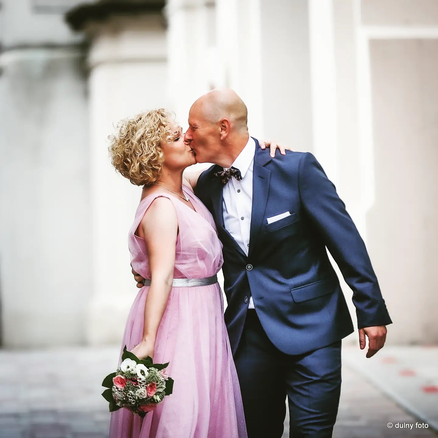 Pocałunek pary młodej: łysy pan w granatowym garniturze z muszką i panna młoda w pudrowej sukni z bukietem na tle białych kolumn.