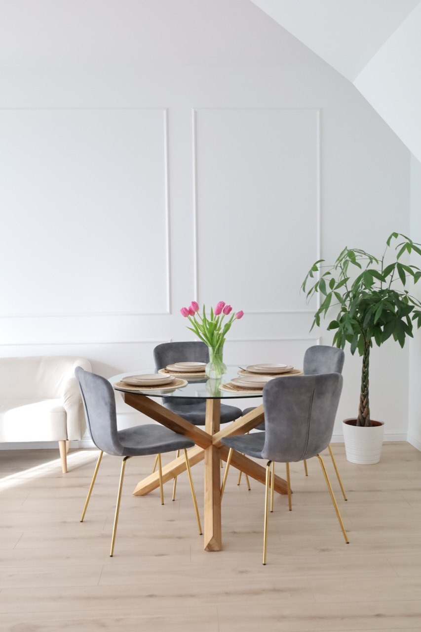 Jasne wnętrze jadalni z okrągłym, szklanym stołem na drewnianej podstawie, szare krzesła z złotymi nogami, różowe tulipany w wazonie i duża roślina doniczkowa, dekoracyjne panele ścienne.
