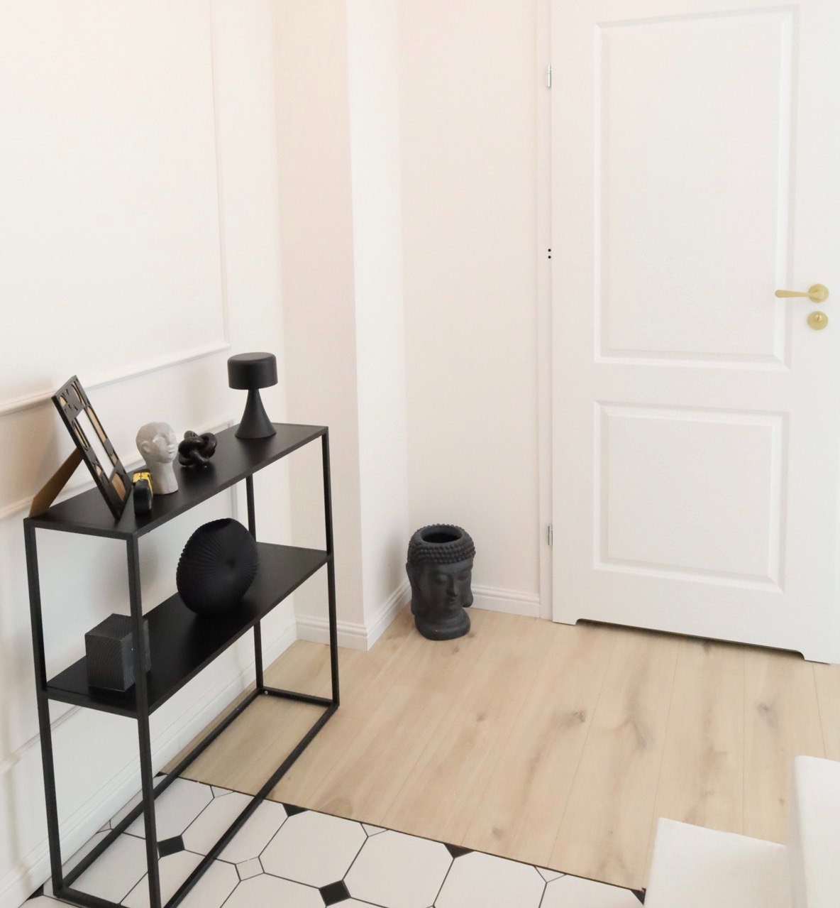 Minimalistyczne wnętrze: czarny, metalowy regał z dekoracjami (ramka, popiersie, wazon) na tle białej ściany z listwami i białych drzwi z złotą klamką, podłoga wyłożona panelami i fragmentem...
