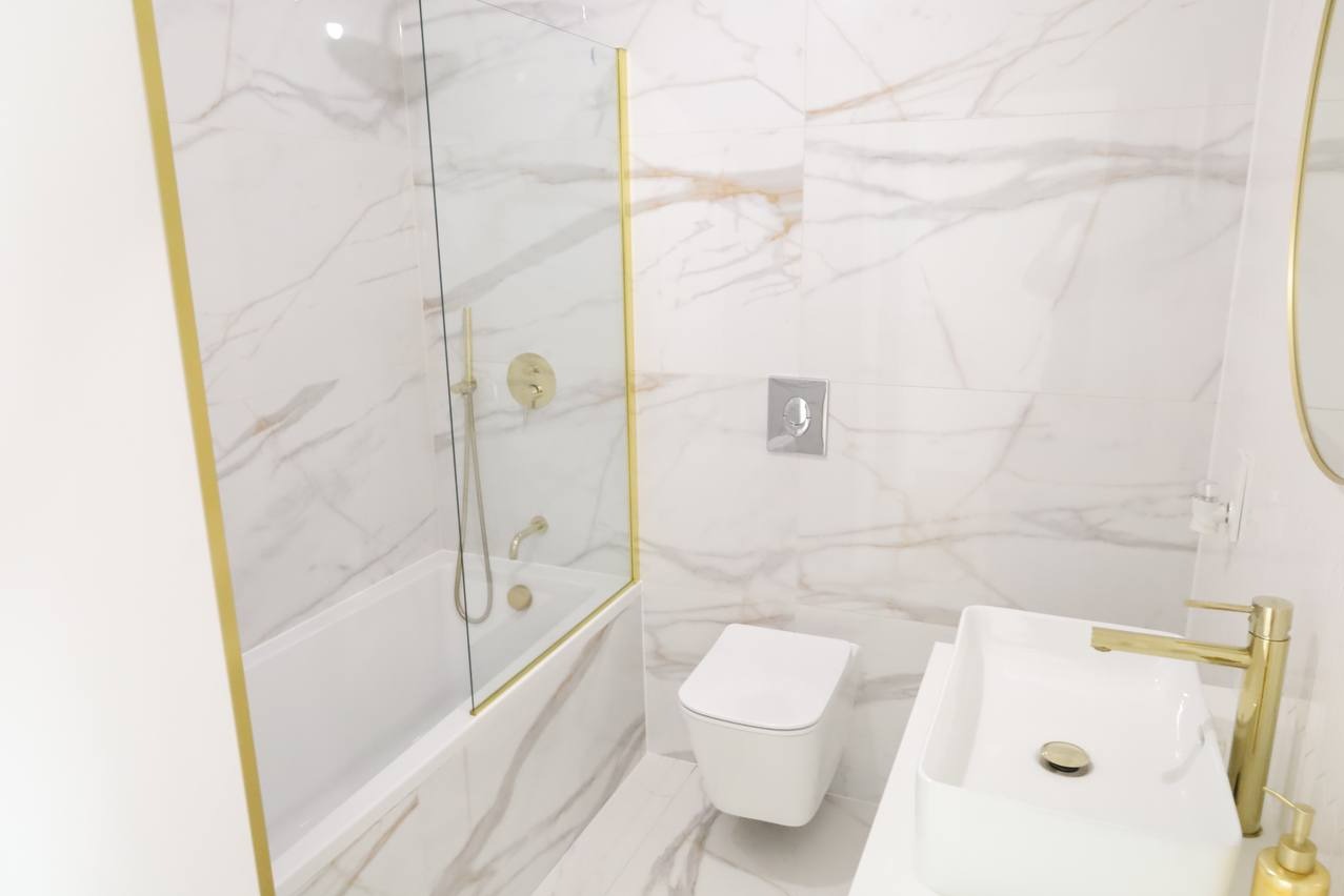 Elegancka, biała łazienka z marmurowymi płytkami, złotymi akcentami na armaturze, kabinie prysznicowej i lustrze, oraz nowoczesną toaletą i umywalką.