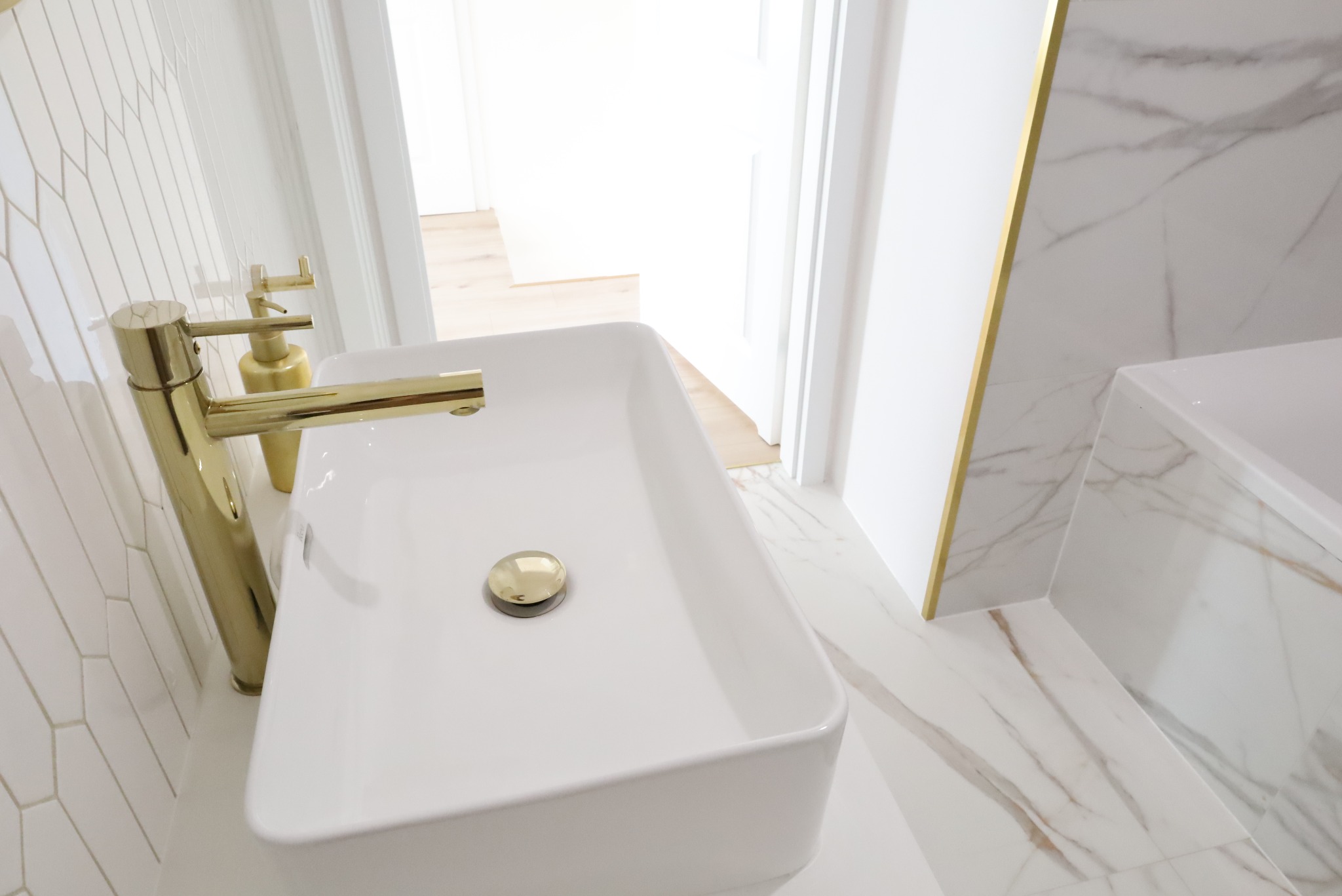 Biała, prostokątna umywalka z pozłacaną armaturą i odpływem na tle białych płytek w jodełkę i marmurowych płytek podłogowych z żyłami w odcieniach złota.