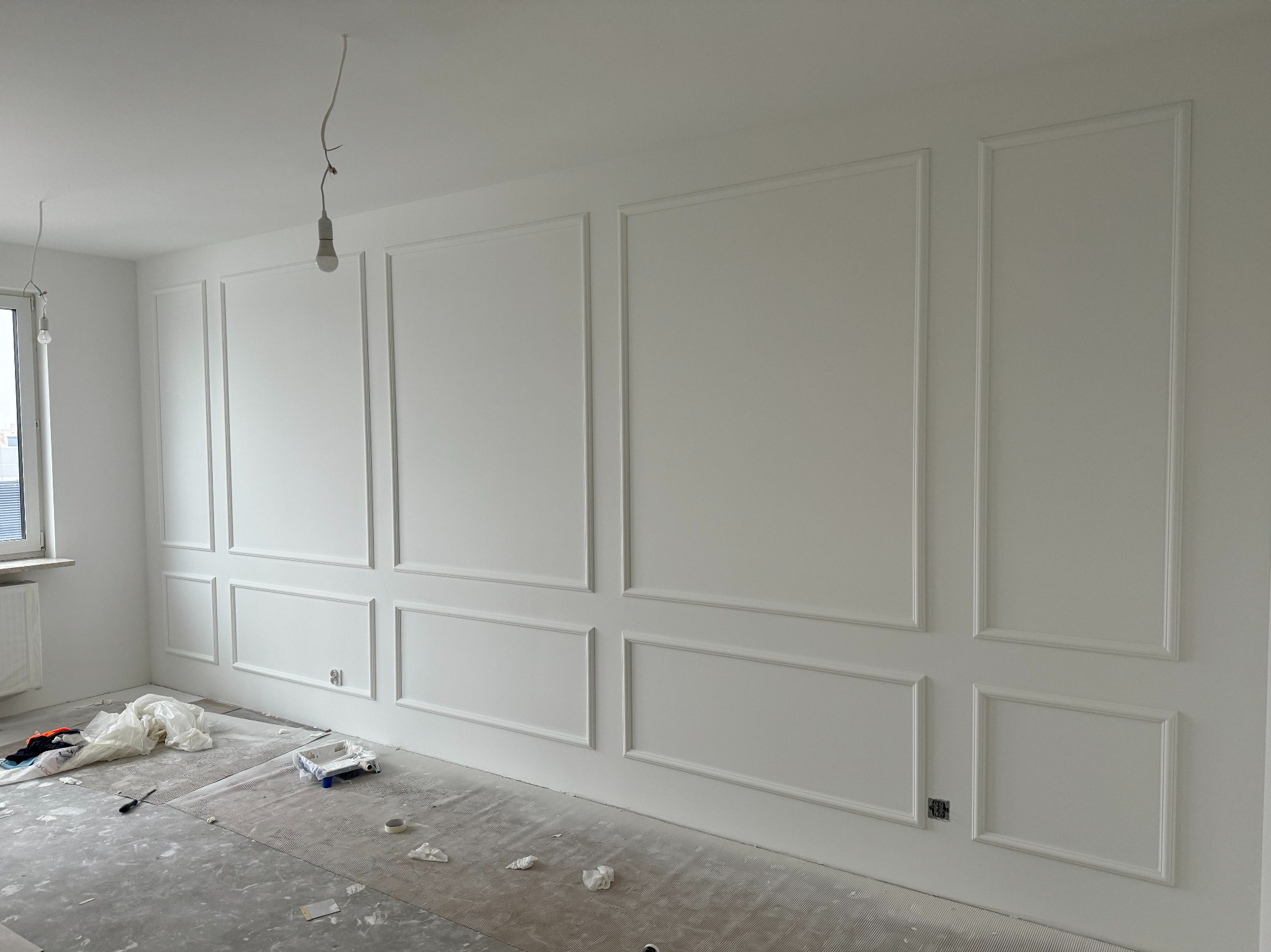 Białe ściany z dekoracyjnymi listwami w stylu angielskim, widoczne gniazdka elektryczne, podłoga zabezpieczona folią malarską, surowa żarówka. Wykończenie wnętrz.