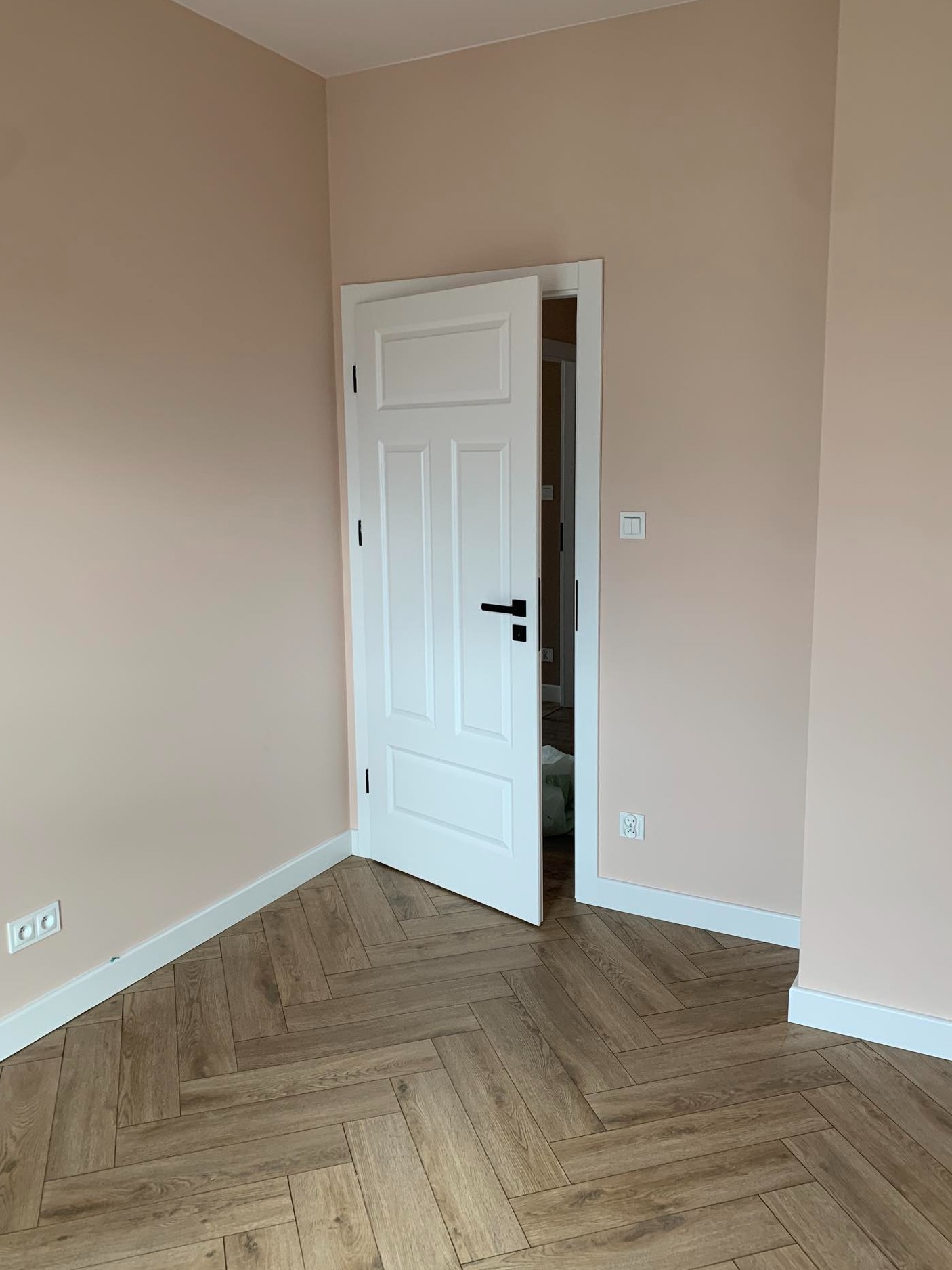 Jasnoróżowe ściany, białe listwy przypodłogowe i białe drzwi z czarną klamką, panele ułożone w jodełkę, widok na wnętrze drugiego pomieszczenia.