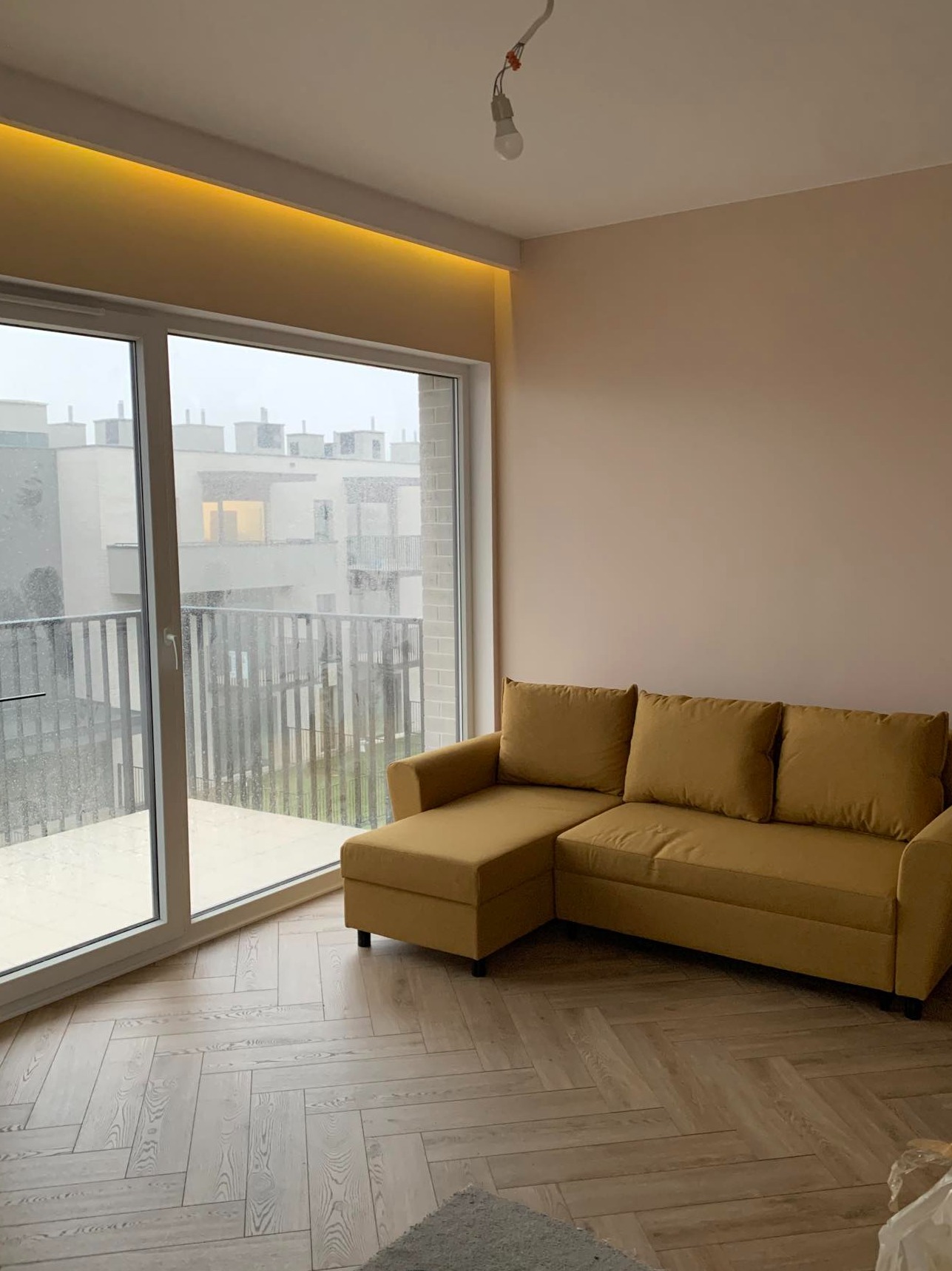 Jasne wnętrze z parkietem w jodełkę i żółtą sofą narożną, widok na balkon i budynki mieszkalne przez duże okno, oświetlenie LED podsufitowe, surowa żarówka zwisająca z sufitu.