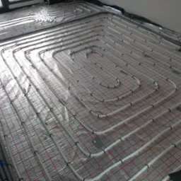 Rozłożone rury ogrzewania podłogowego na folii z siatką, widoczne złącza i kolektor po lewej stronie.