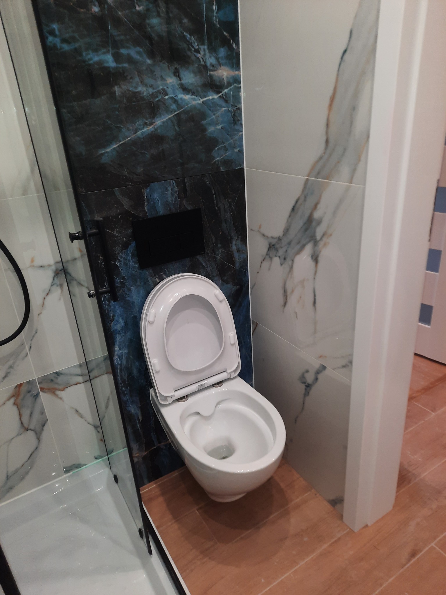 Nowoczesna toaleta wisząca z otwartą klapą, obudowana płytkami imitującymi marmur w odcieniach bieli i granatu, z widoczną kabiną prysznicową w tle i drewnopodobną podłogą.