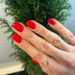 Dłoń z czerwonymi, kwadratowymi paznokciami na tle zielonej rośliny w doniczce, widoczny złoty pierścionek z trzema kamieniami.