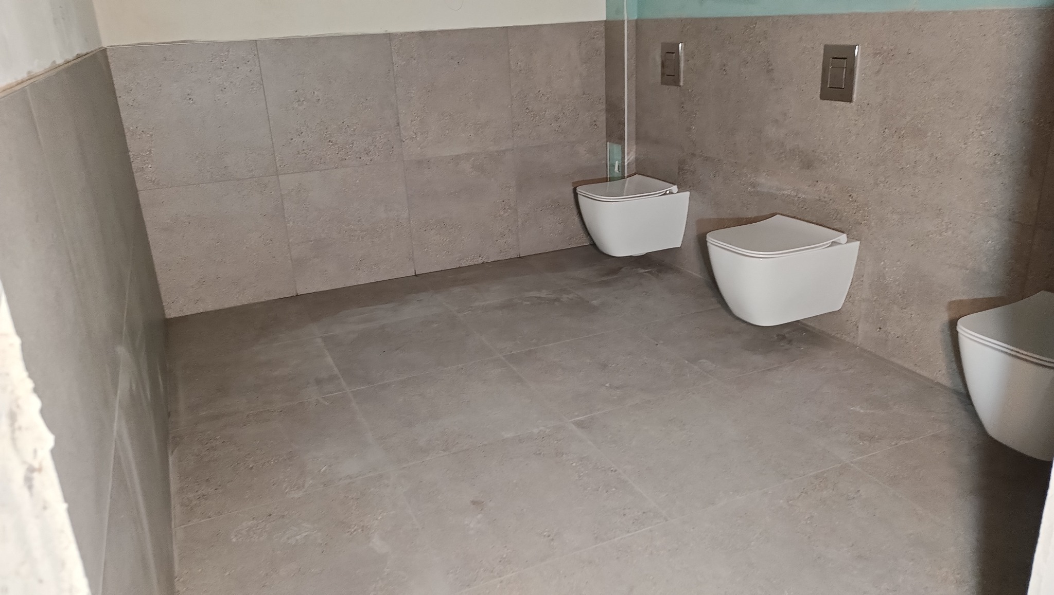 Nowoczesna łazienka w szarych tonacjach z podwieszanymi toaletami i płytkami imitującymi kamień na ścianach i podłodze. Minimalistyczny design.